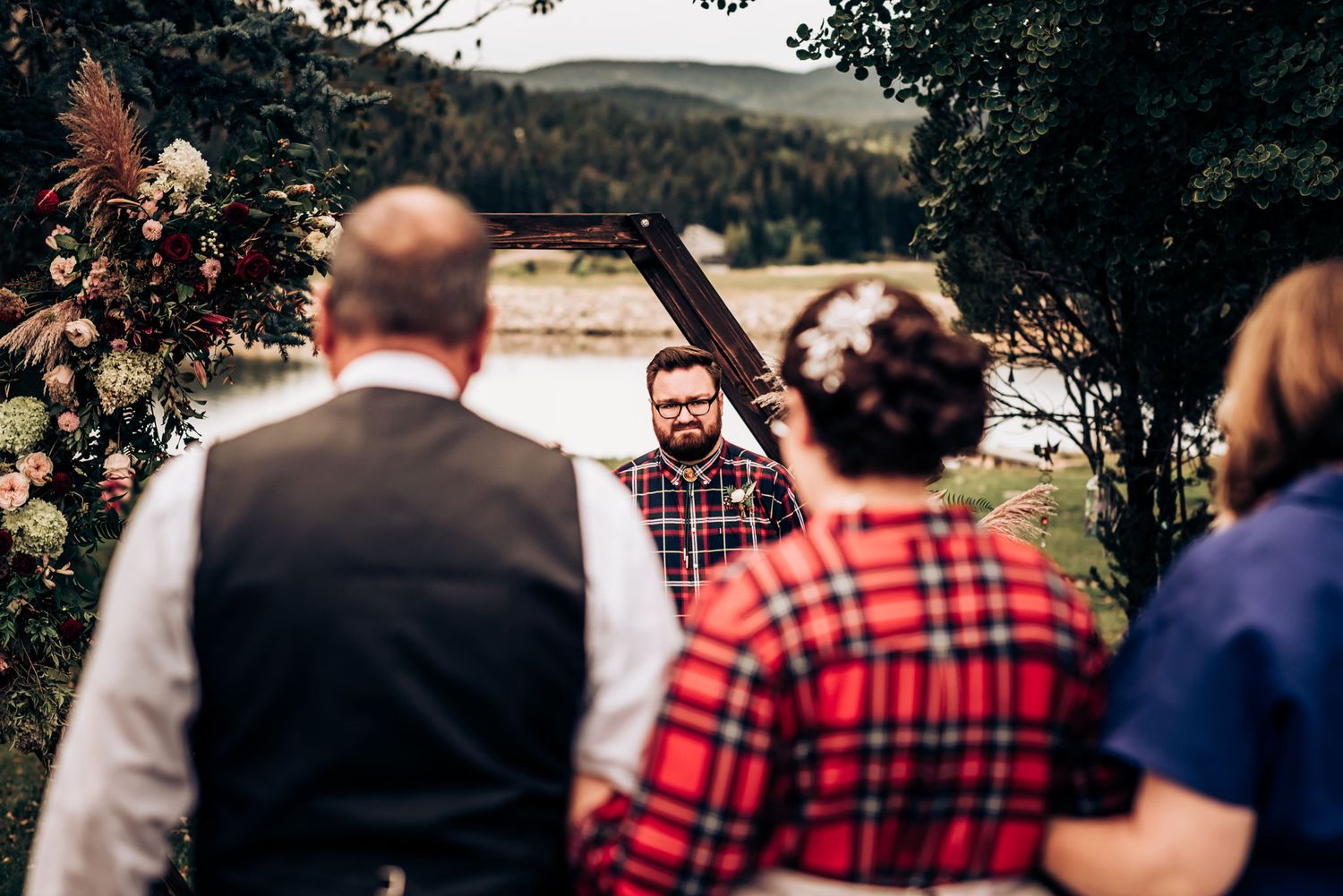 An outdoor wedding ceremony takes place in front of a rustic stone wall with floral decorations and mountain views.