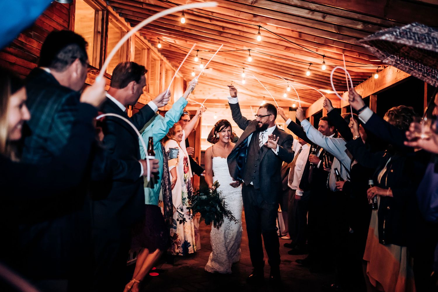 Wedding guests raise their arms in celebration under wooden beams with warm lighting.