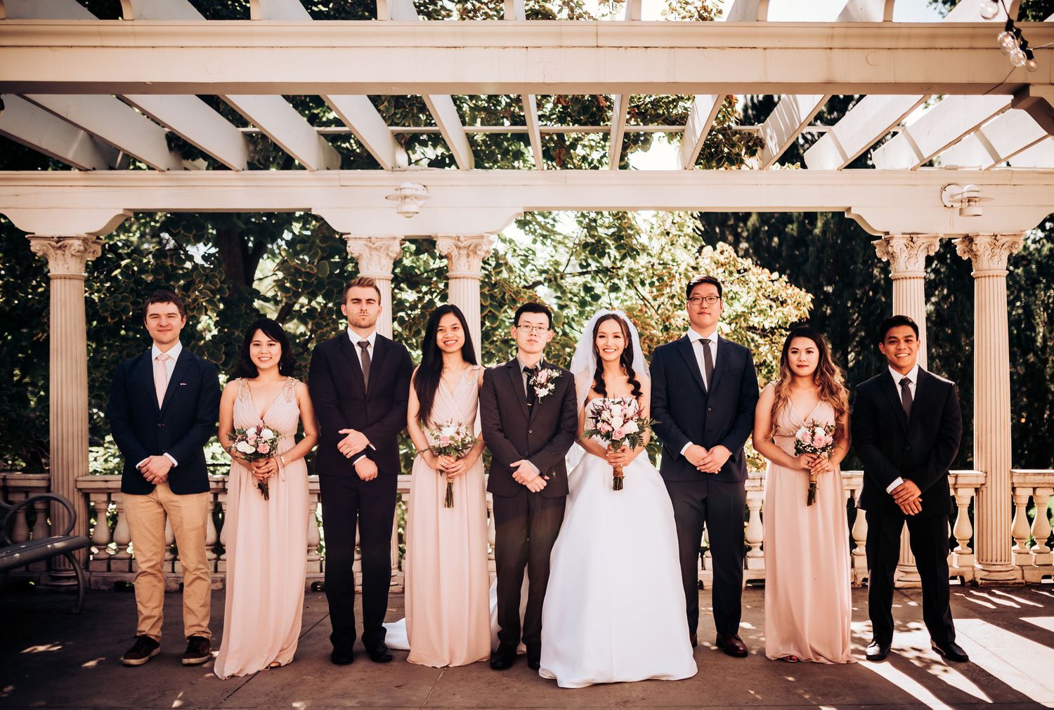 Wedding party poses under white pergola with bridesmaids in blush gowns.