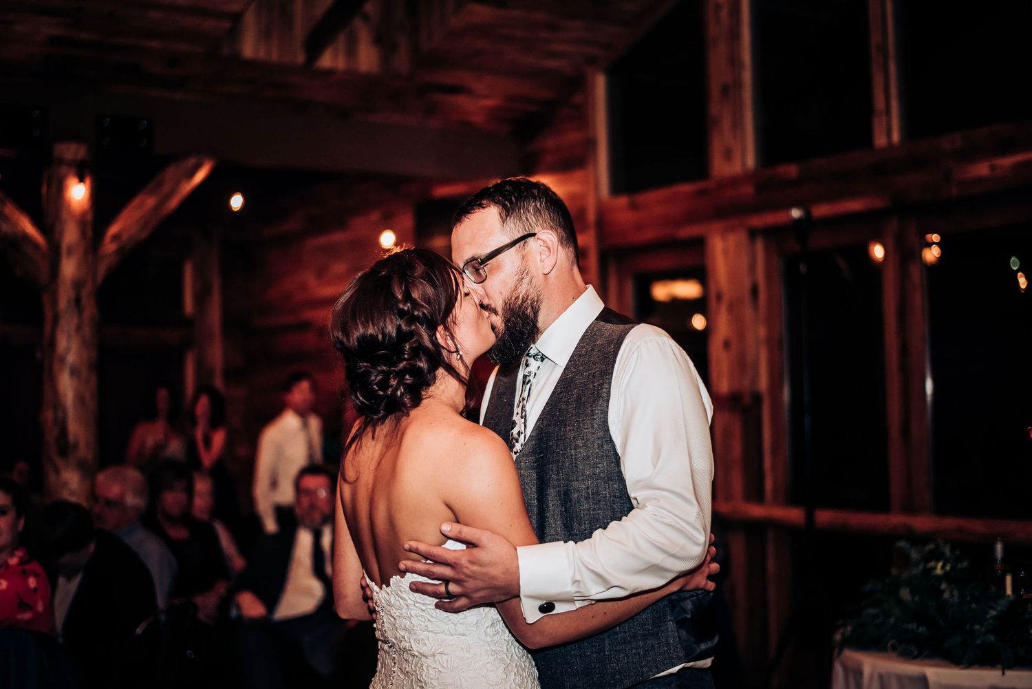 Intimate dance moment in a warmly lit rustic wooden venue with guests in background.