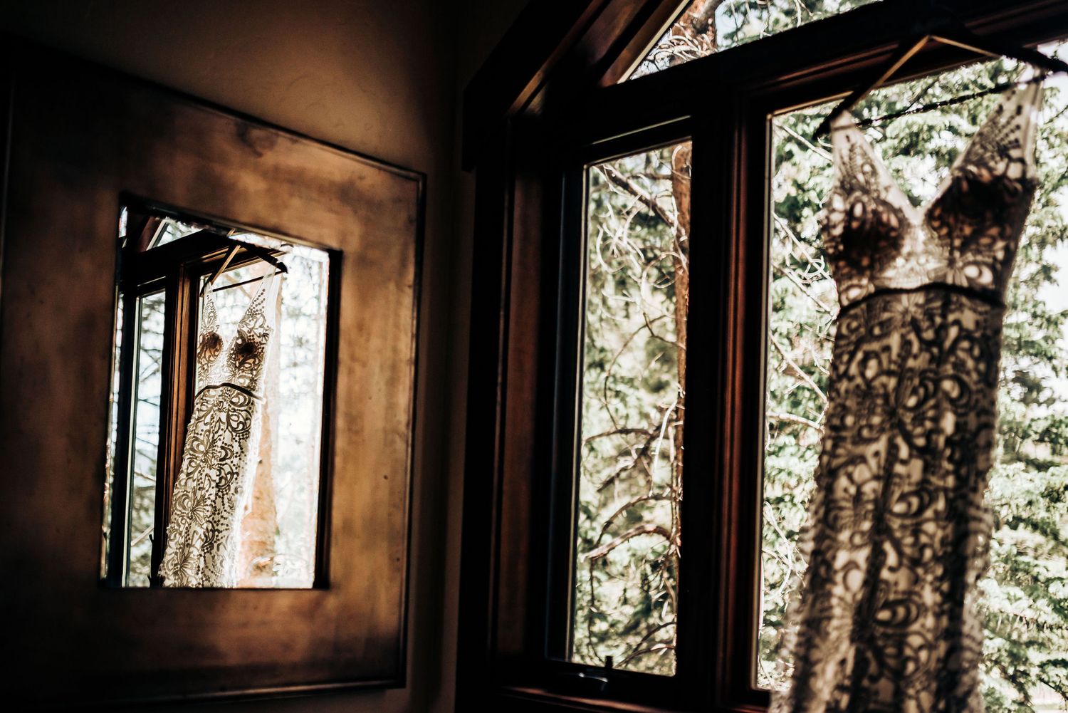 Wedding dress hanging in window with natural light creating artistic reflection.