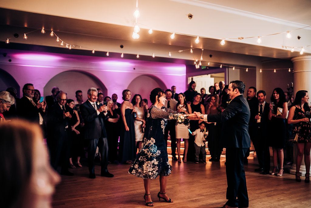 Wedding guests gather on the dance floor during a reception with purple mood lighting and string lights.