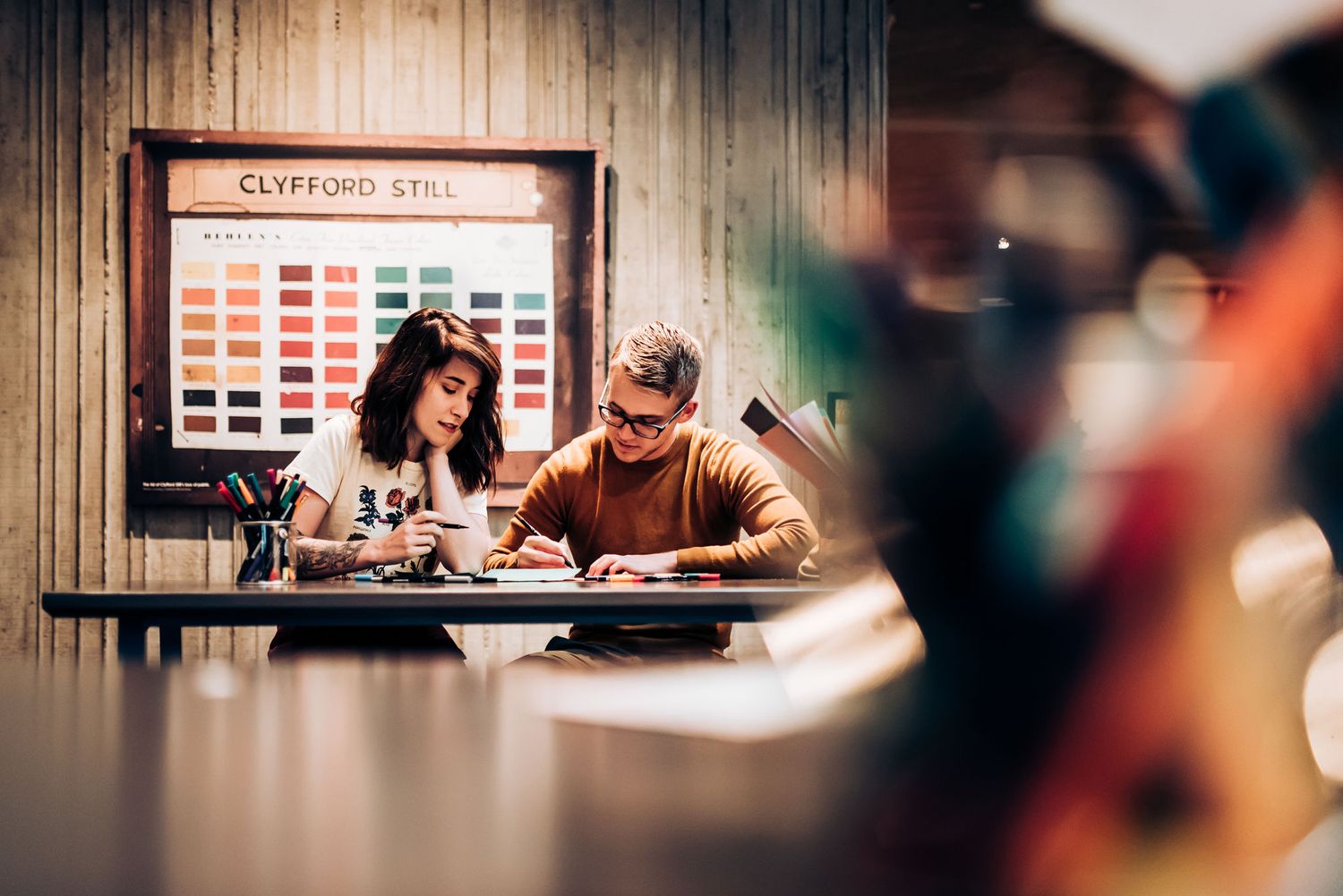 Two artists work on a project at a desk with color swatches displayed on the wall behind them.