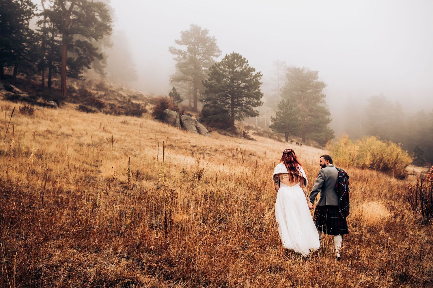 Scenic mountain wedding photo with couple walking through misty autumn field.