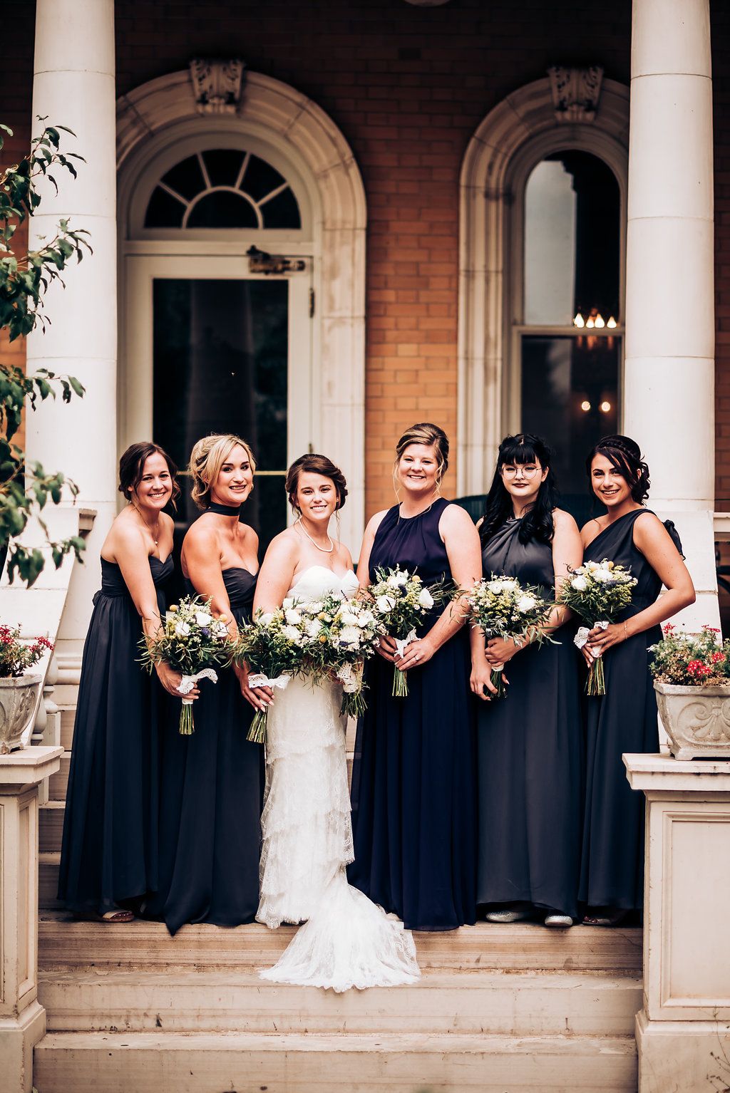 Bridal party in long navy dresses poses formally on stone steps of historic building.
