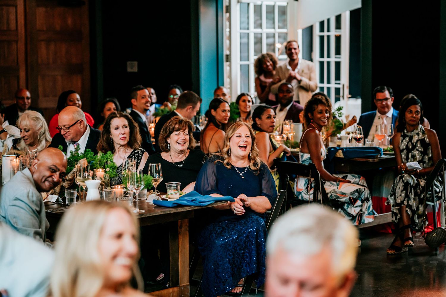 Wedding guests laugh and smile during a reception celebration at tables filled with drinks and decorations.