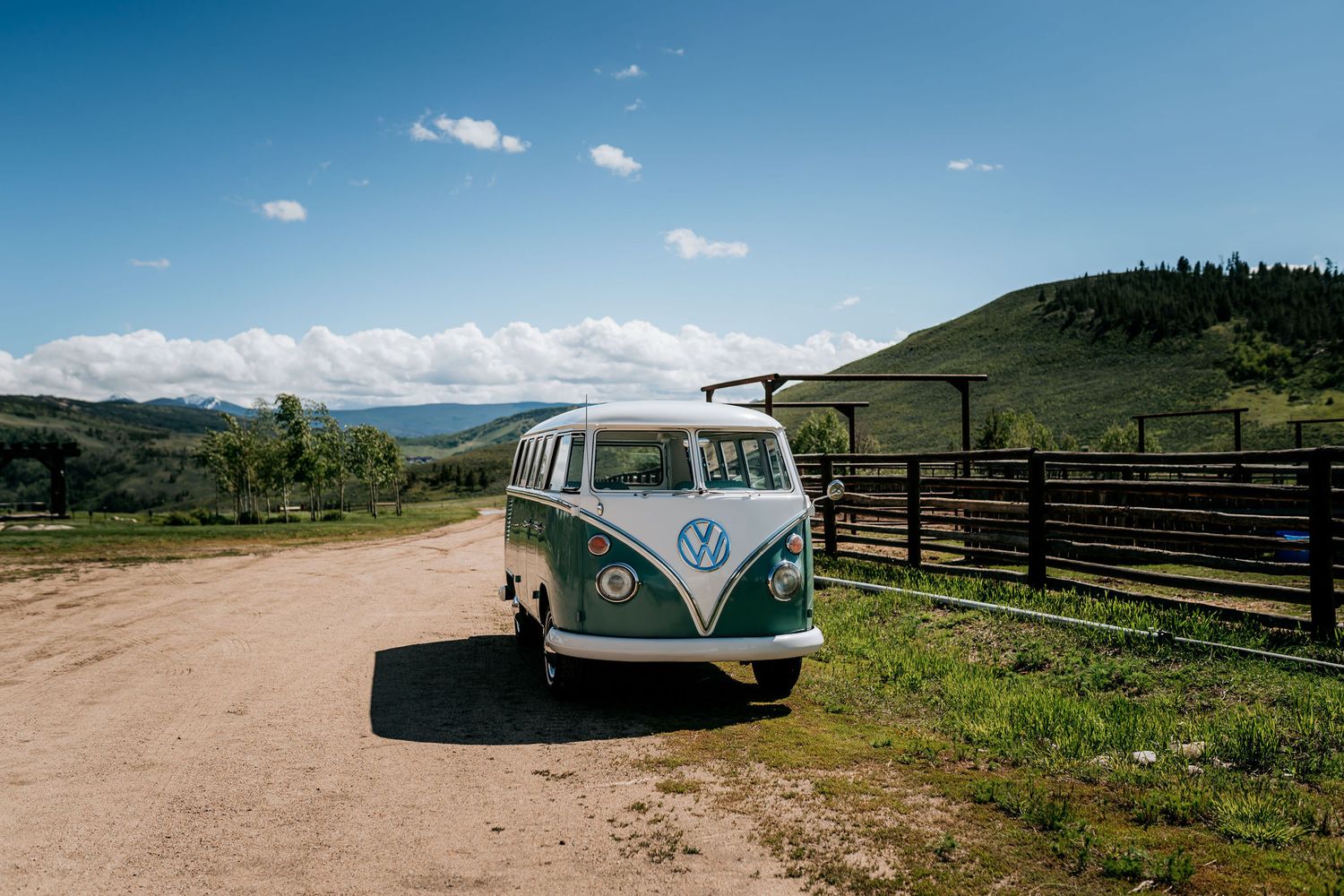 A classic Volkswagen Type 2 bus parked on a dirt road against scenic mountain backdrop with blue skies.