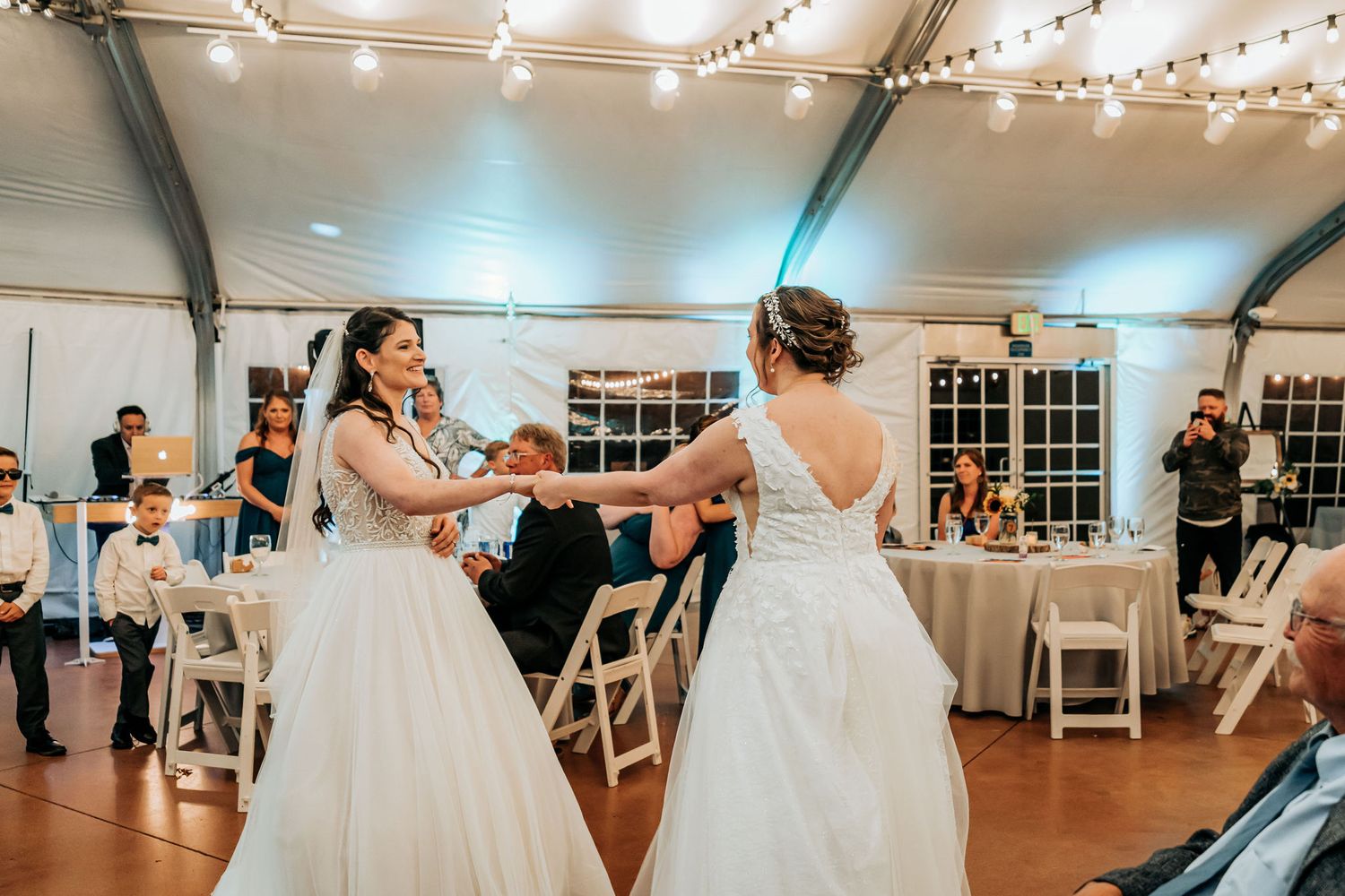 Two people in white gowns dancing together under string lights at a wedding reception.