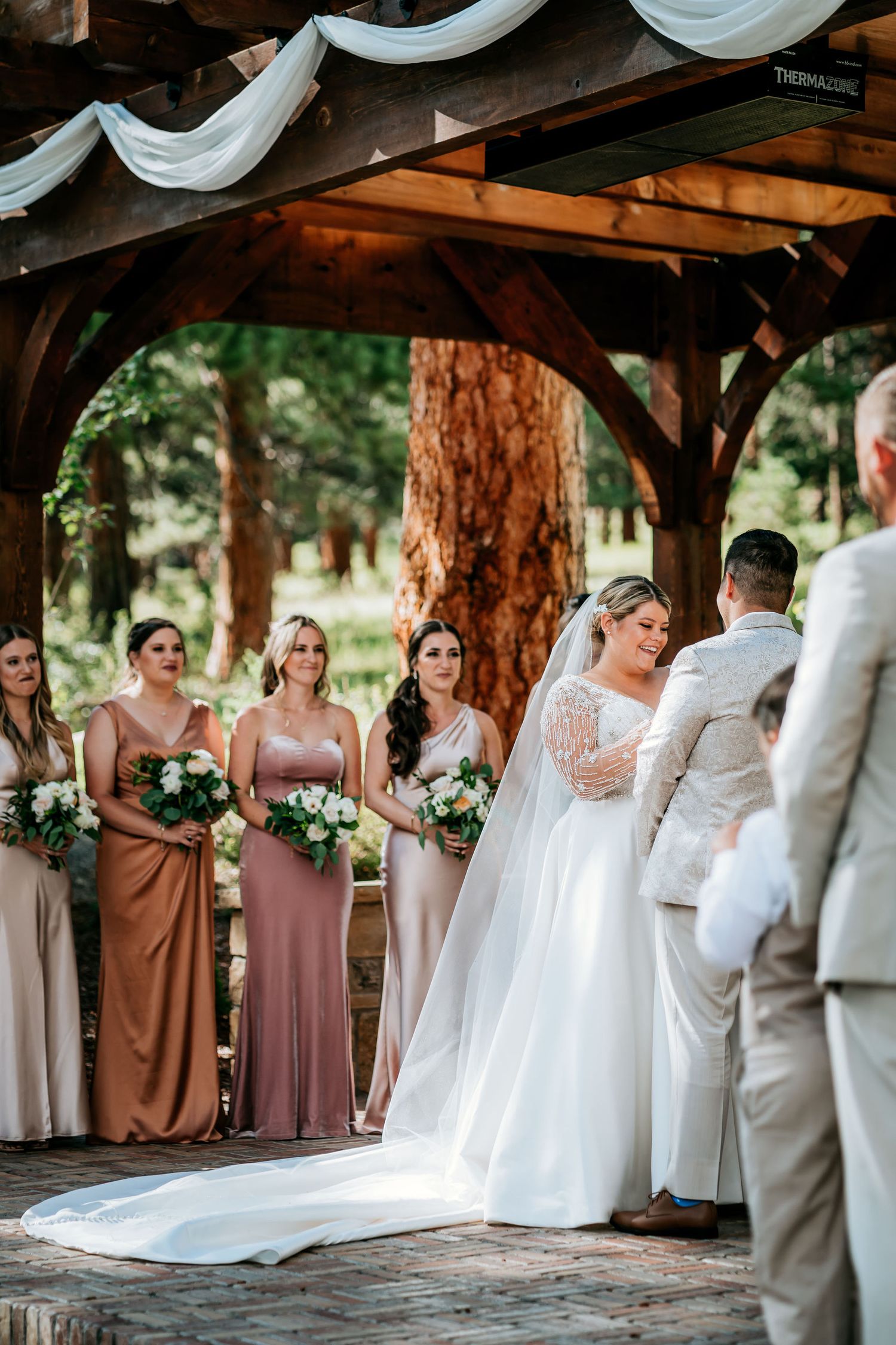 Wedding ceremony under a rustic wooden pergola with draped white fabric and bridesmaids in blush dresses.