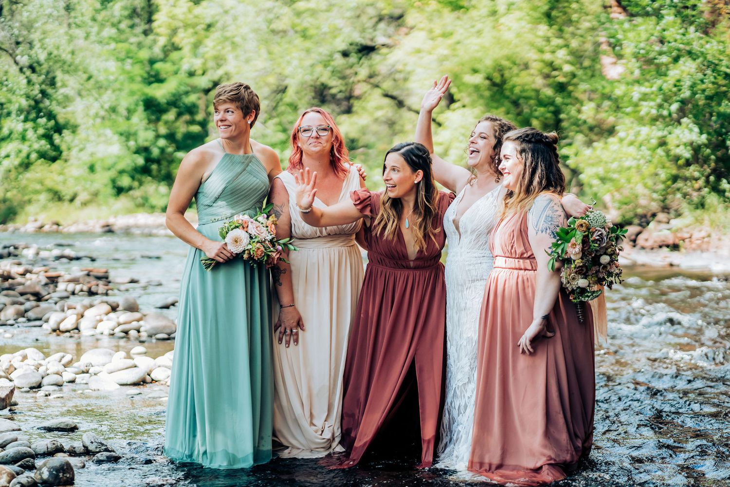 Bridesmaids in pastel dresses pose together by rocky riverbed.