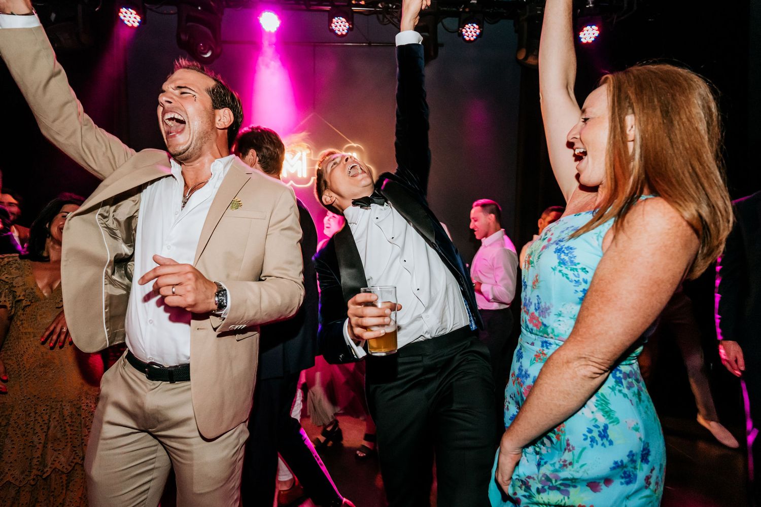 Party guests dancing enthusiastically with hands raised under colorful lighting at indoor venue.