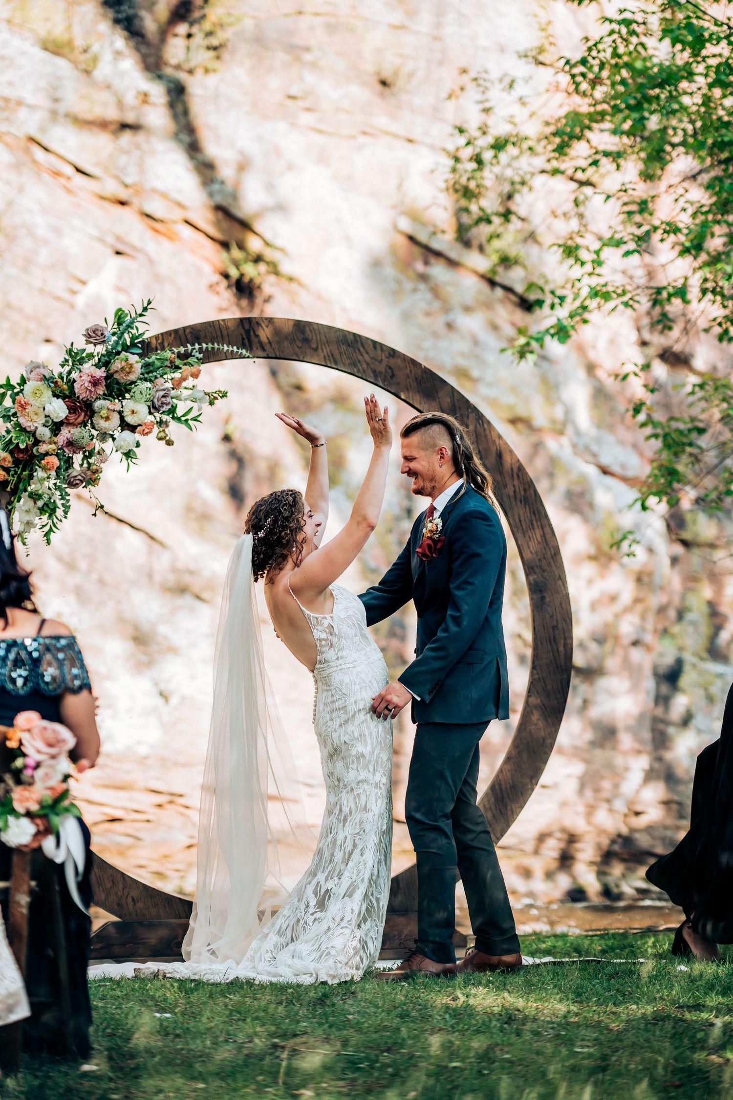 Outdoor wedding ceremony features couple standing beneath circular floral arch against rocky cliff backdrop.