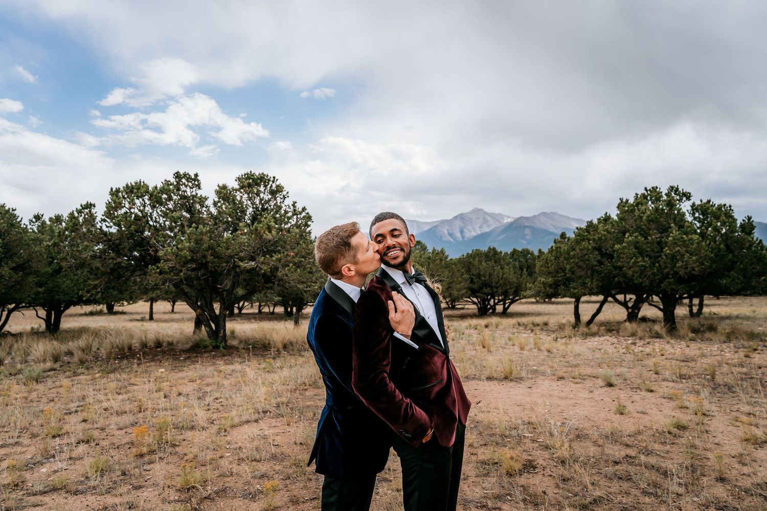 A couple embraces in an outdoor setting with mountain peaks and scattered trees in the background.