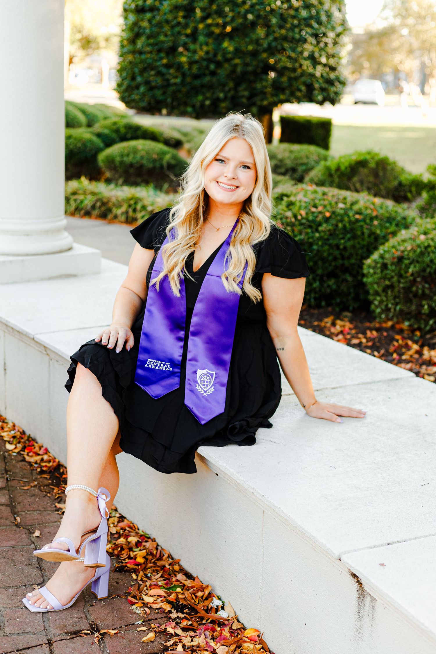 Graduate in black dress and purple stole poses near white columns with autumn leaves.