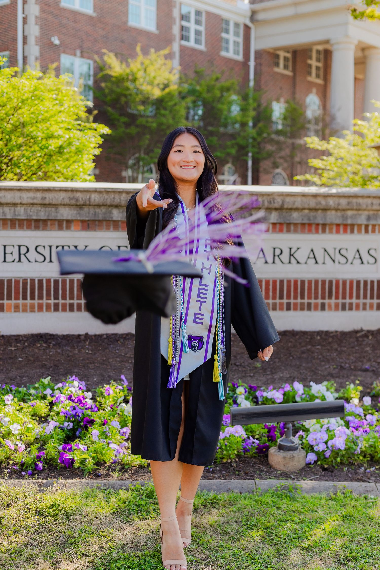 A graduate in academic regalia poses with University of Arkansas signage in front of blooming spring flowers.