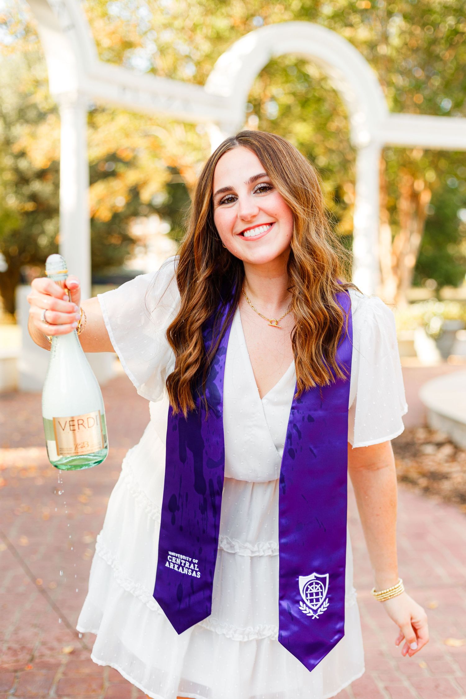 Graduate in white dress and purple stole celebrates with champagne bottle outdoors.