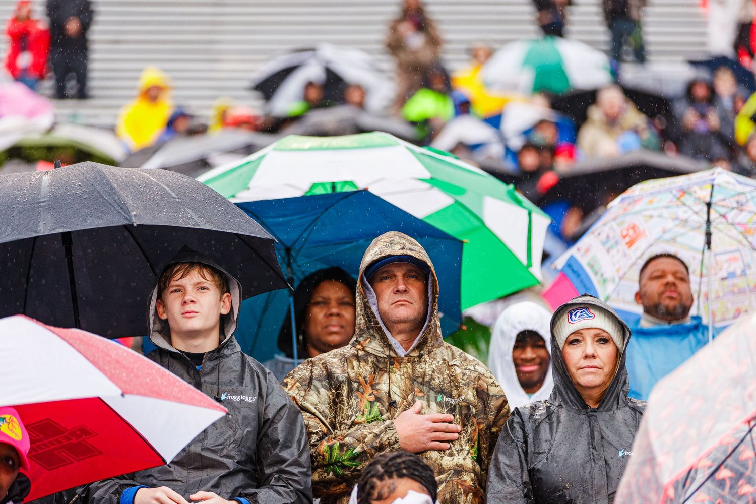 Crowd of spectators huddle under umbrellas during rainy outdoor sporting event.