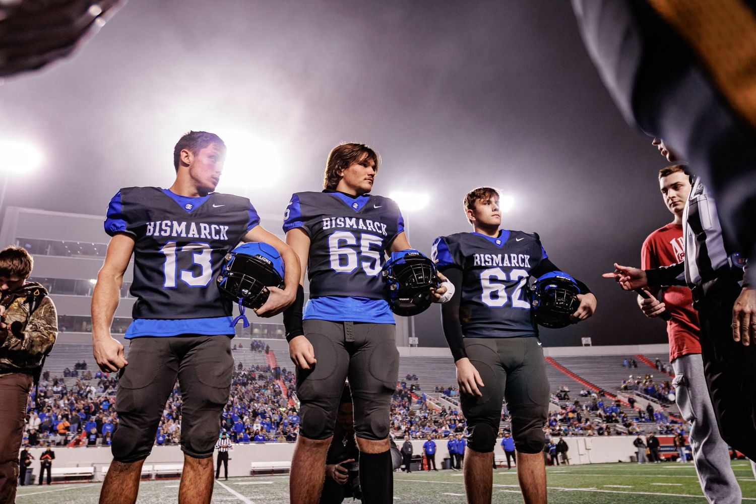 Team members stand together on the football field sideline during a nighttime game.