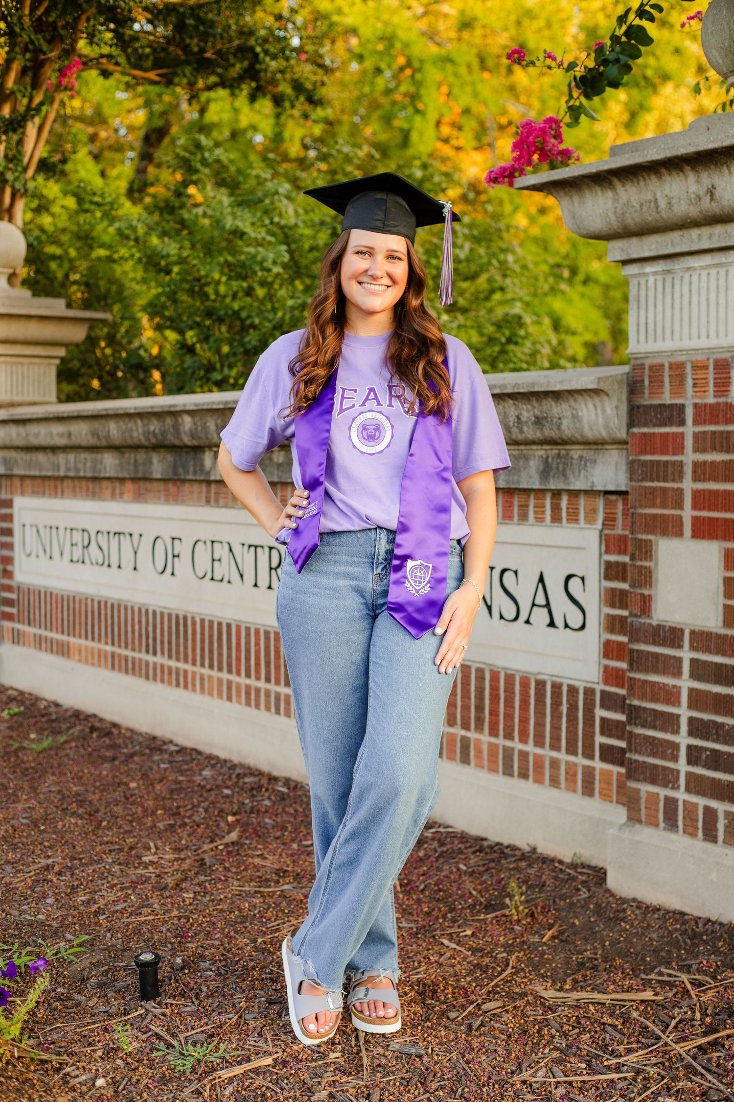 Graduate in cap and purple stole poses by University of Central Arkansas brick wall during fall season.
