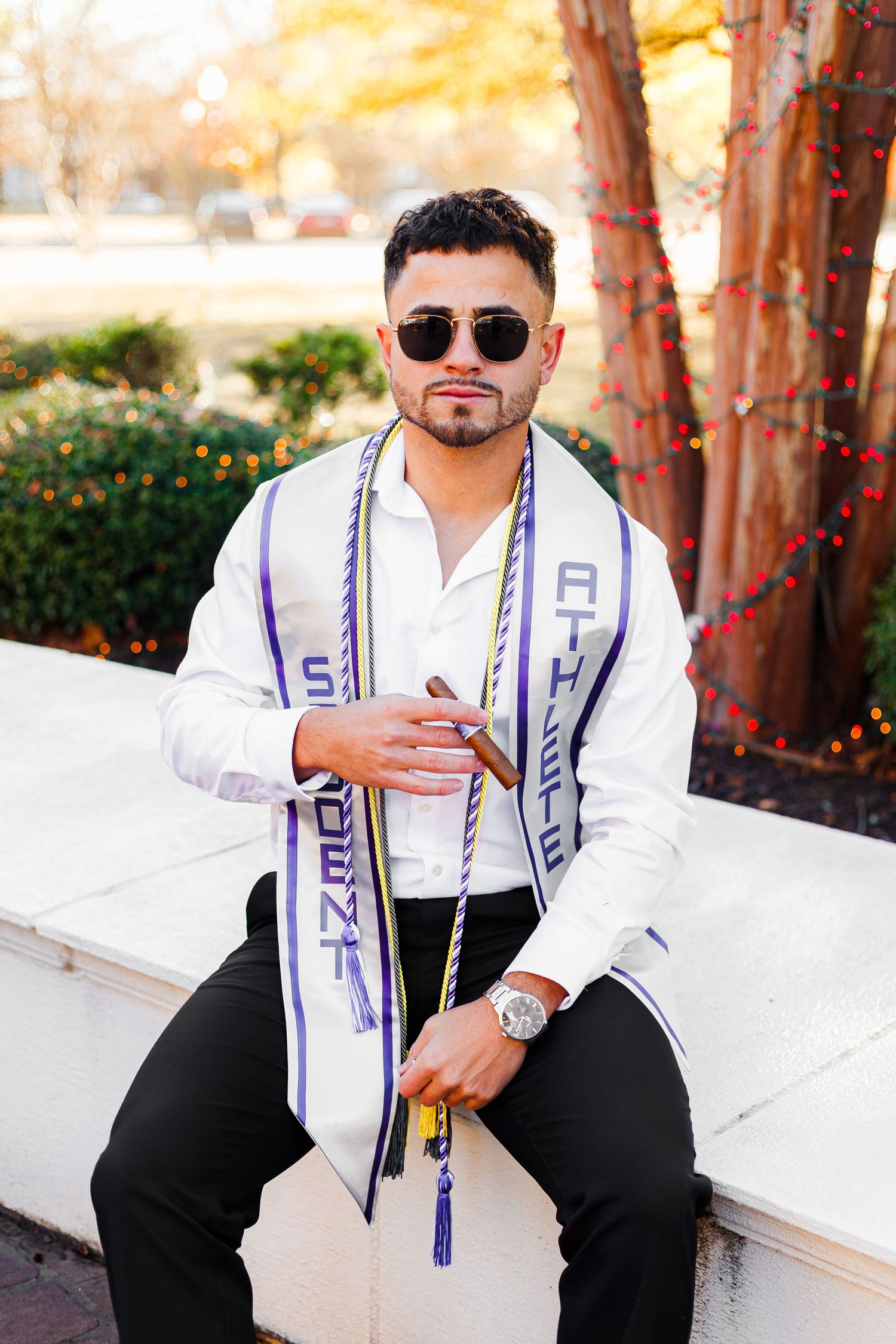 Graduate wearing sunglasses and graduation stole poses on white ledge with decorative lights.