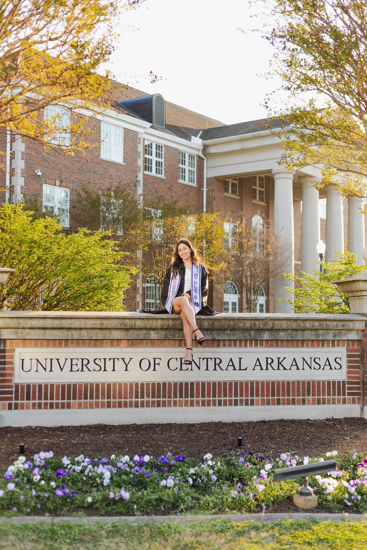 Entrance sign for University of Central Arkansas with brick wall, white columns, and purple flowers blooming in spring.