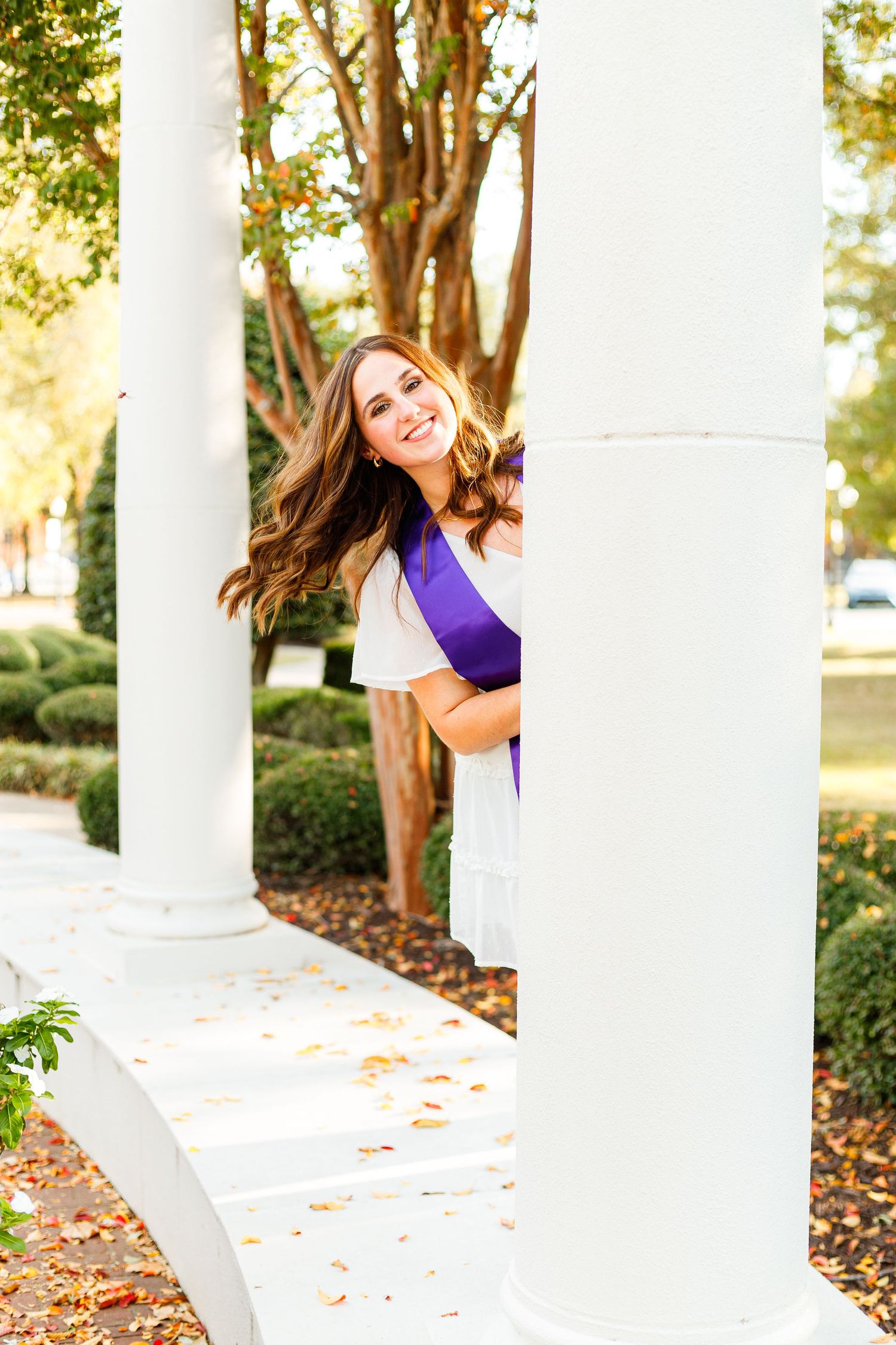 Graduate poses between white columns with fallen autumn leaves on ground.
