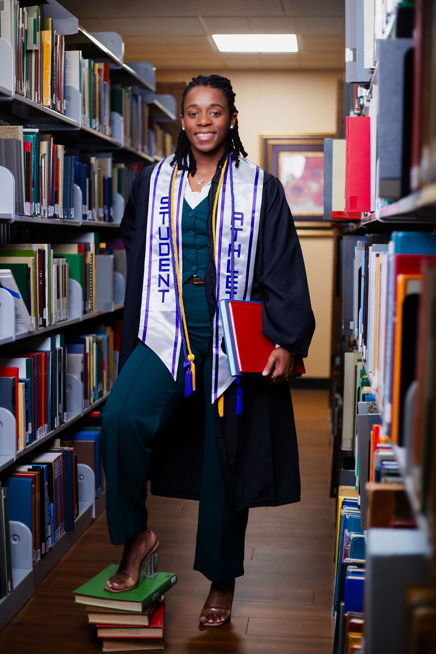 A student in navy graduation robes and ceremonial stole poses for multiple shots between library bookshelves.