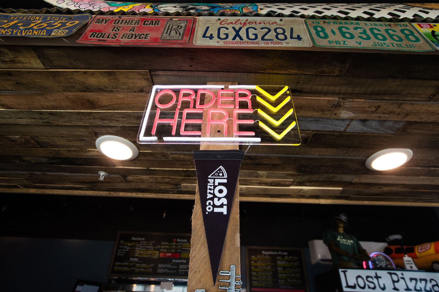 Order here neon sign mounted on wooden beam with license plates in background.