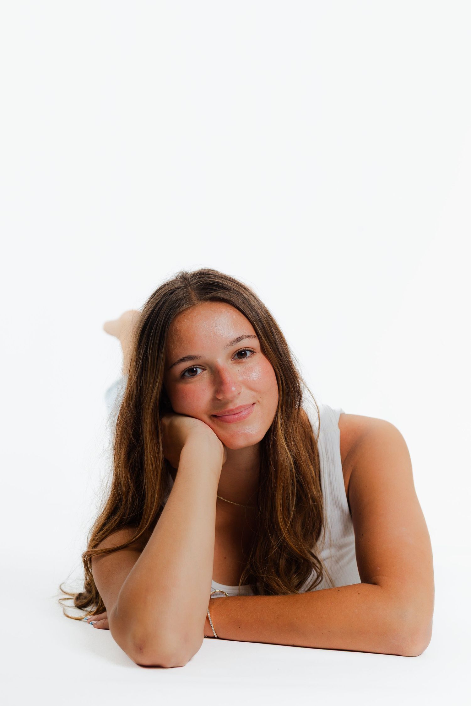 Person with long brown hair wearing a white sleeveless top rests head on hand while laying on white surface.