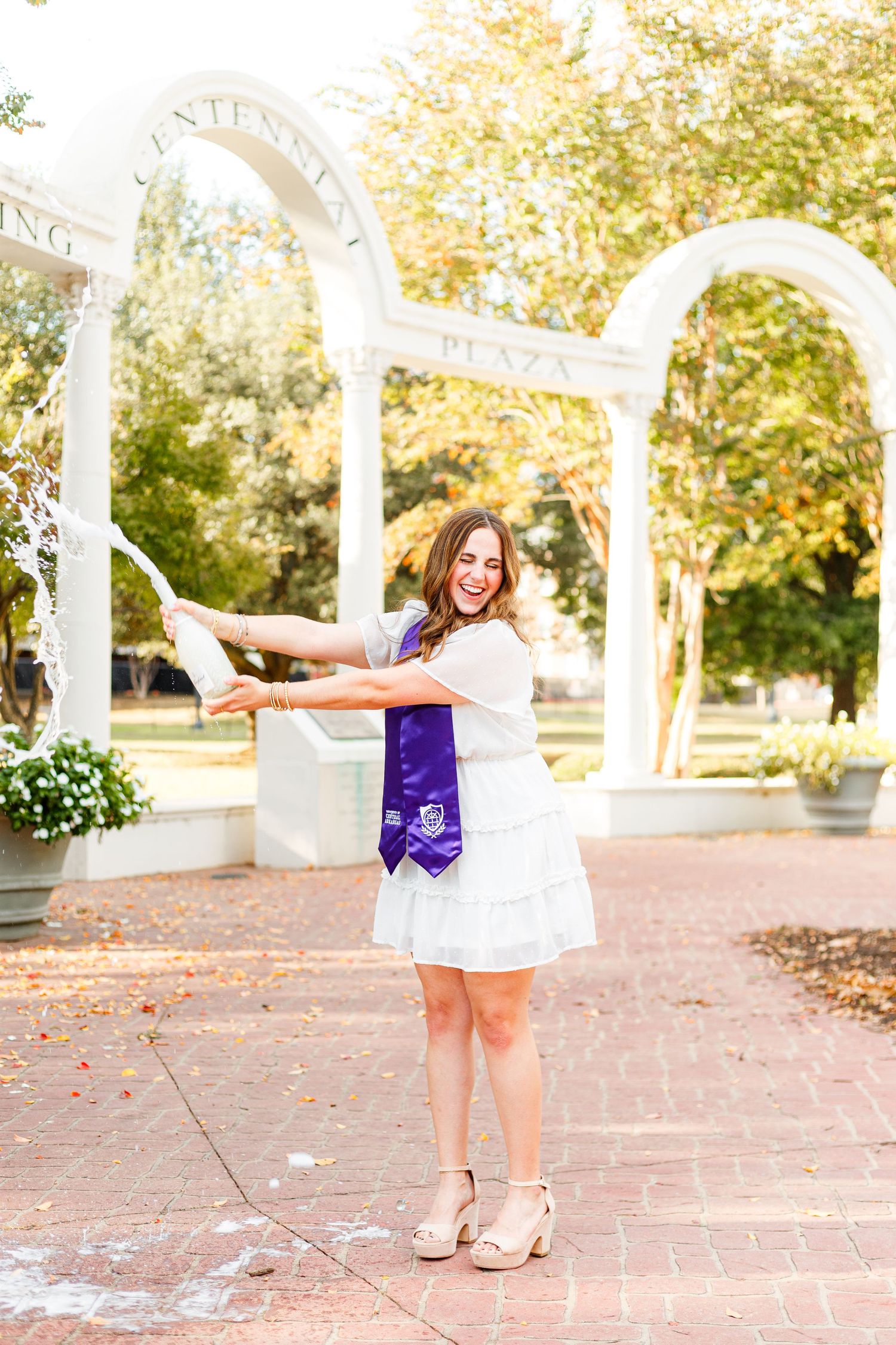Graduate in white dress poses joyfully under white archways on brick pathway.