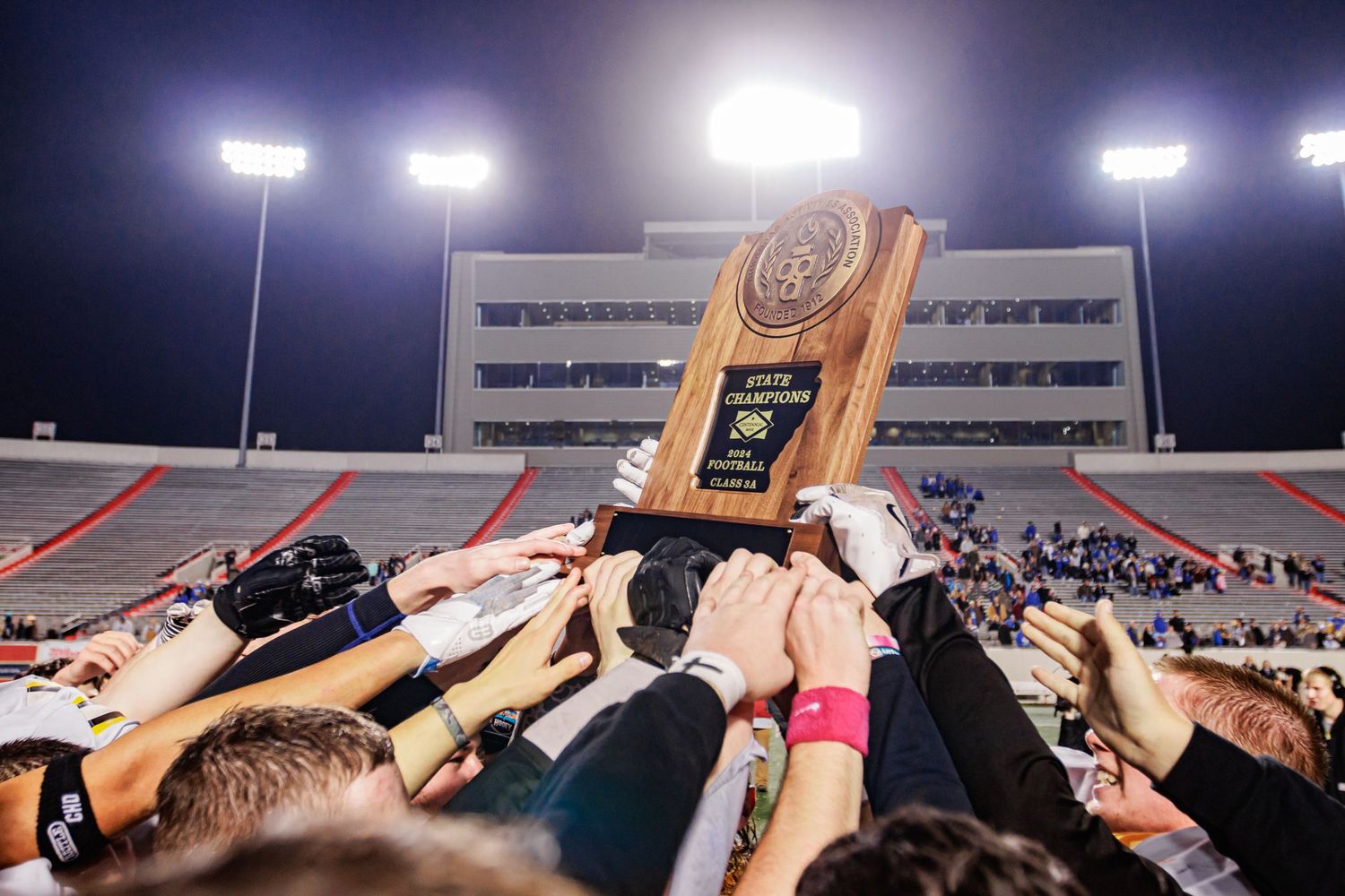 Stadium lights illuminate crowd lifting trophy in celebration at night game.