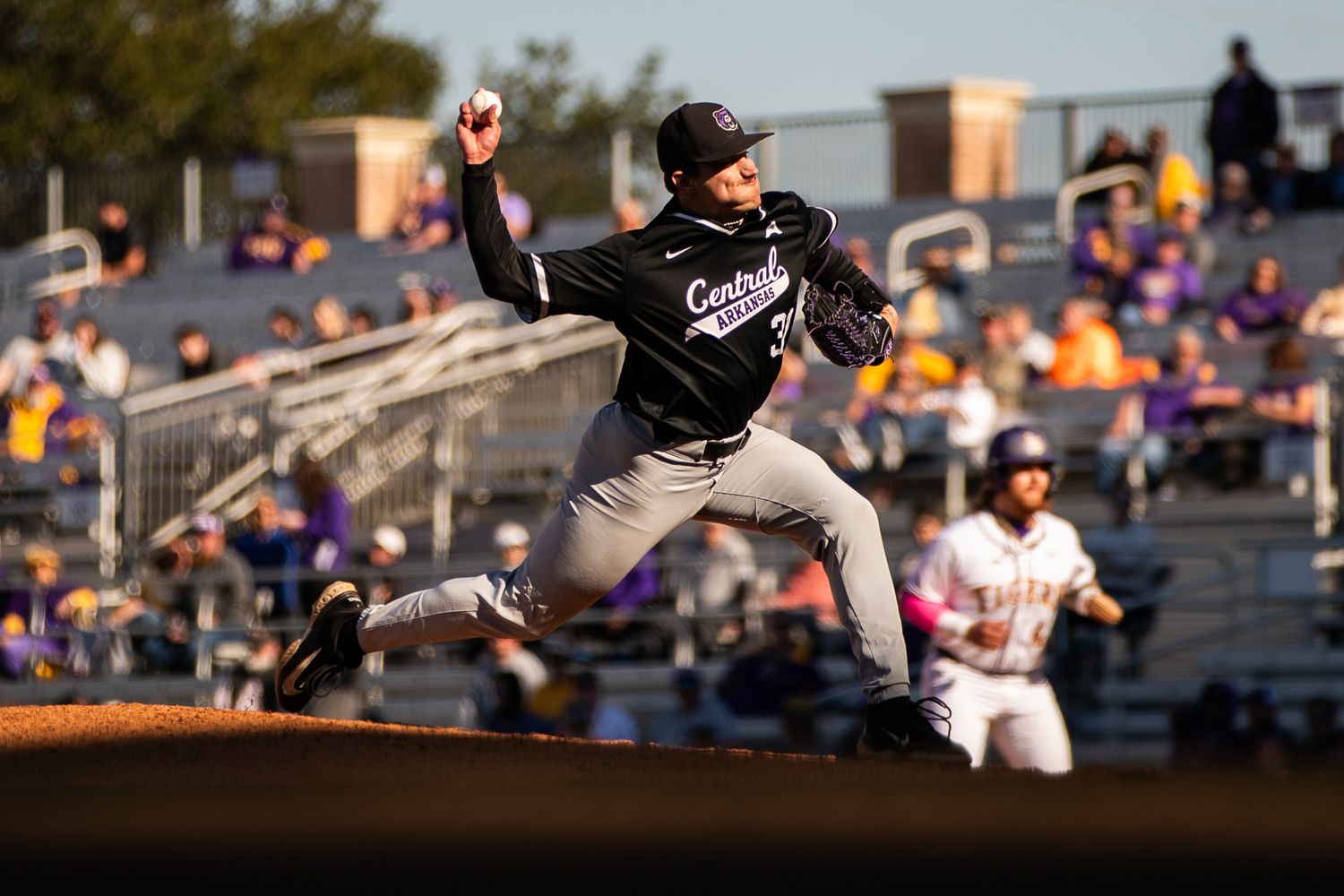 Baseball pitcher captured mid-throw during sunset game with crowd in background.