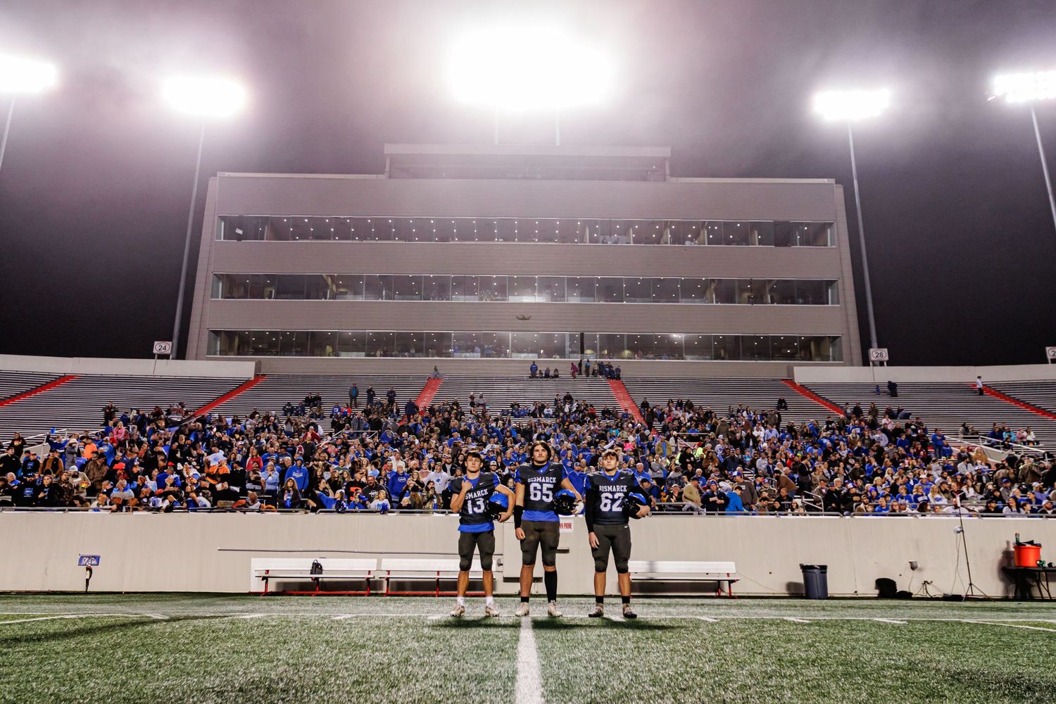 Wide view of stadium field and stands during nighttime football game with bright lights.