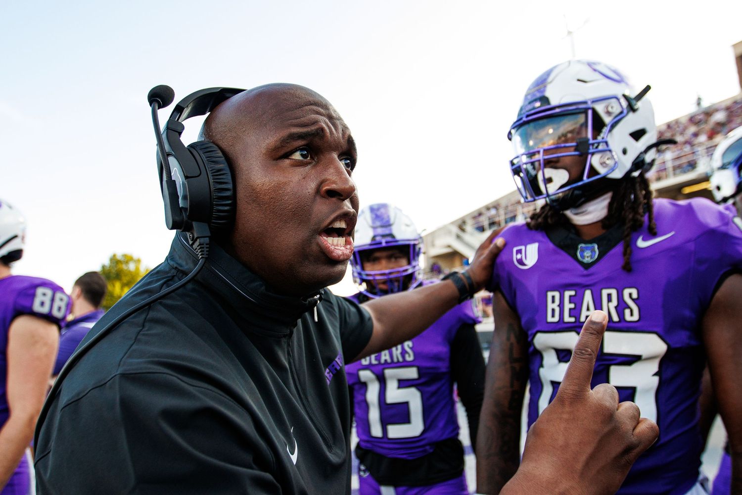 Coach wearing headphones speaks passionately on sideline during college football game.