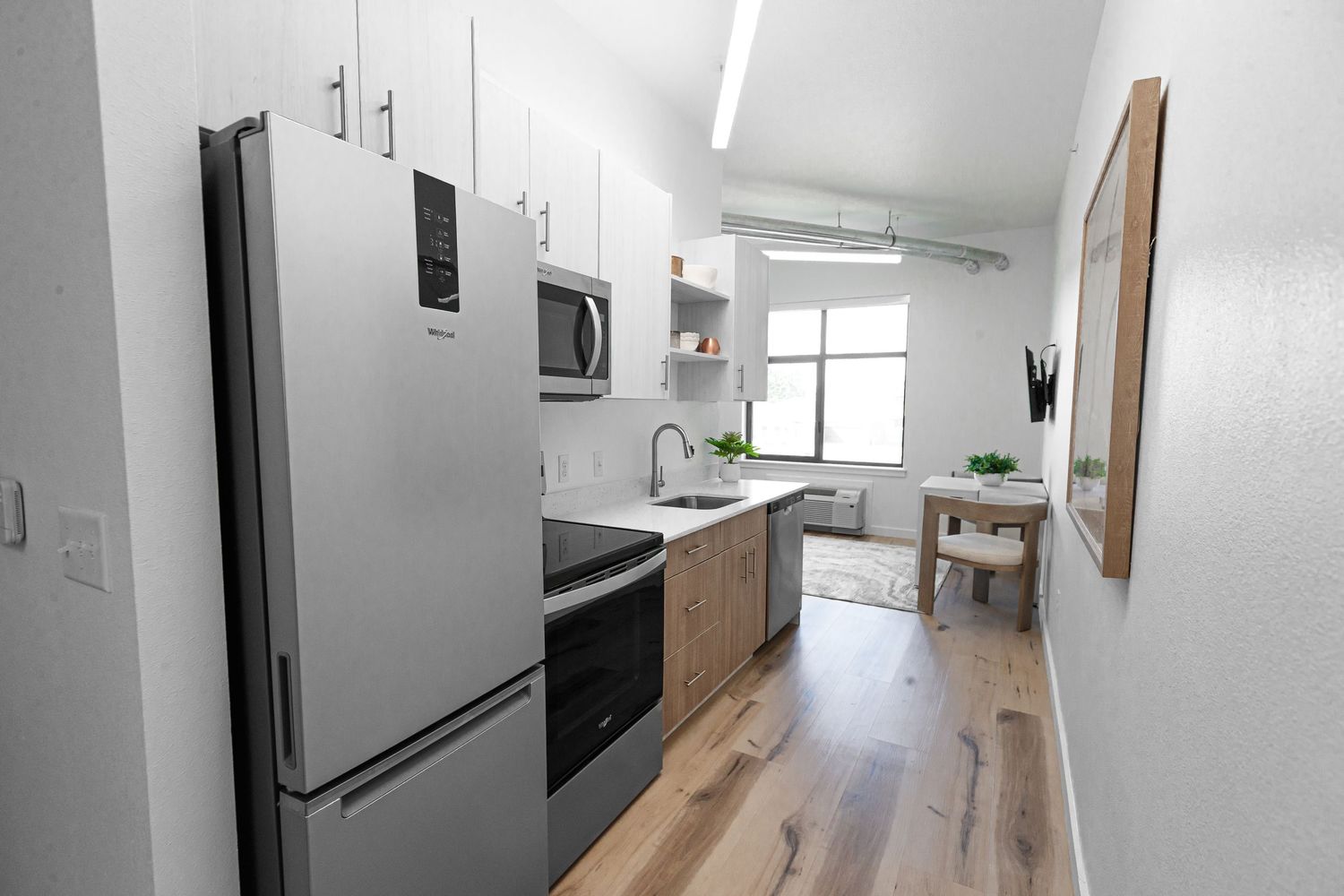 Modern galley kitchen with stainless steel appliances, light wood floors, and white cabinets in bright open layout space.