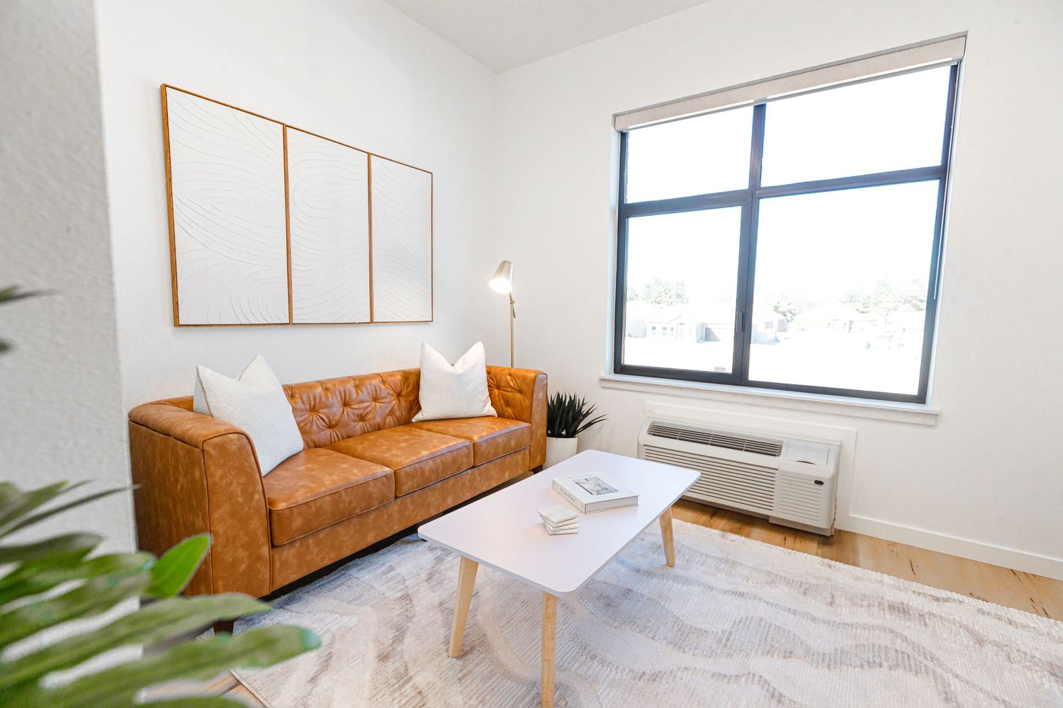 Bright living room with tan leather couch, wooden frames, and large window with natural light.