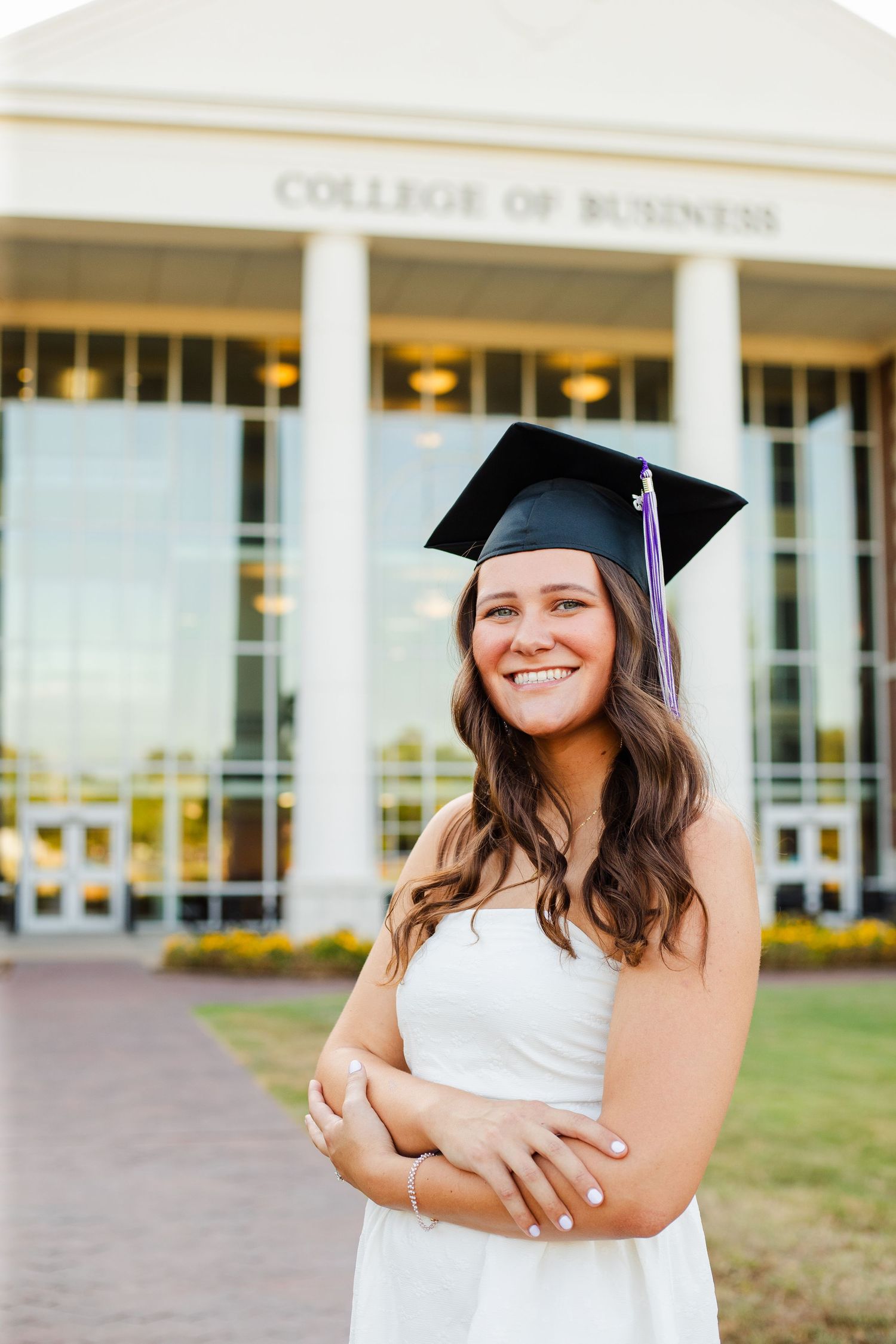 A graduate in a white dress and mortarboard cap poses outside the College of Business building.
