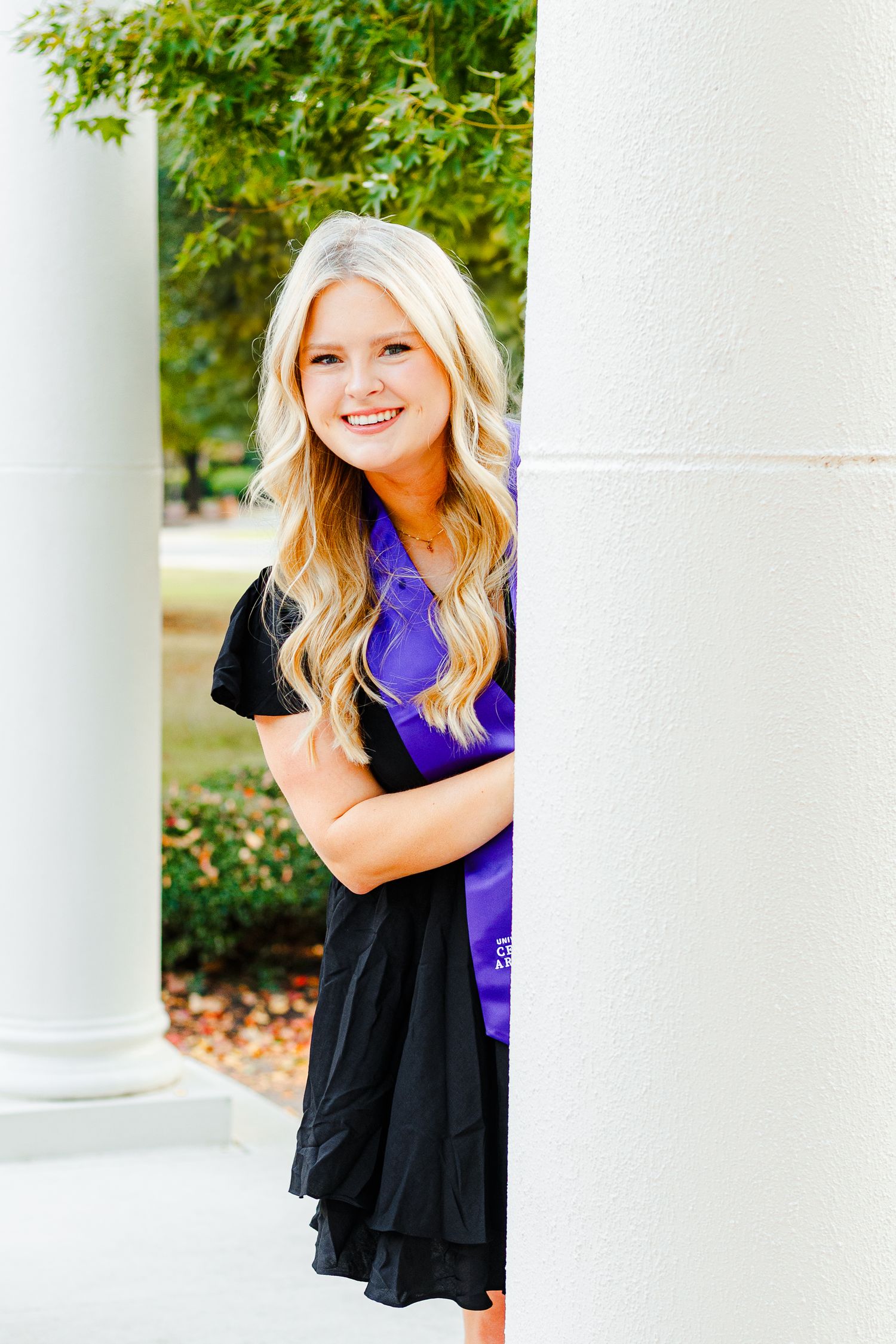 Graduate in black dress with purple sash poses between white architectural columns.