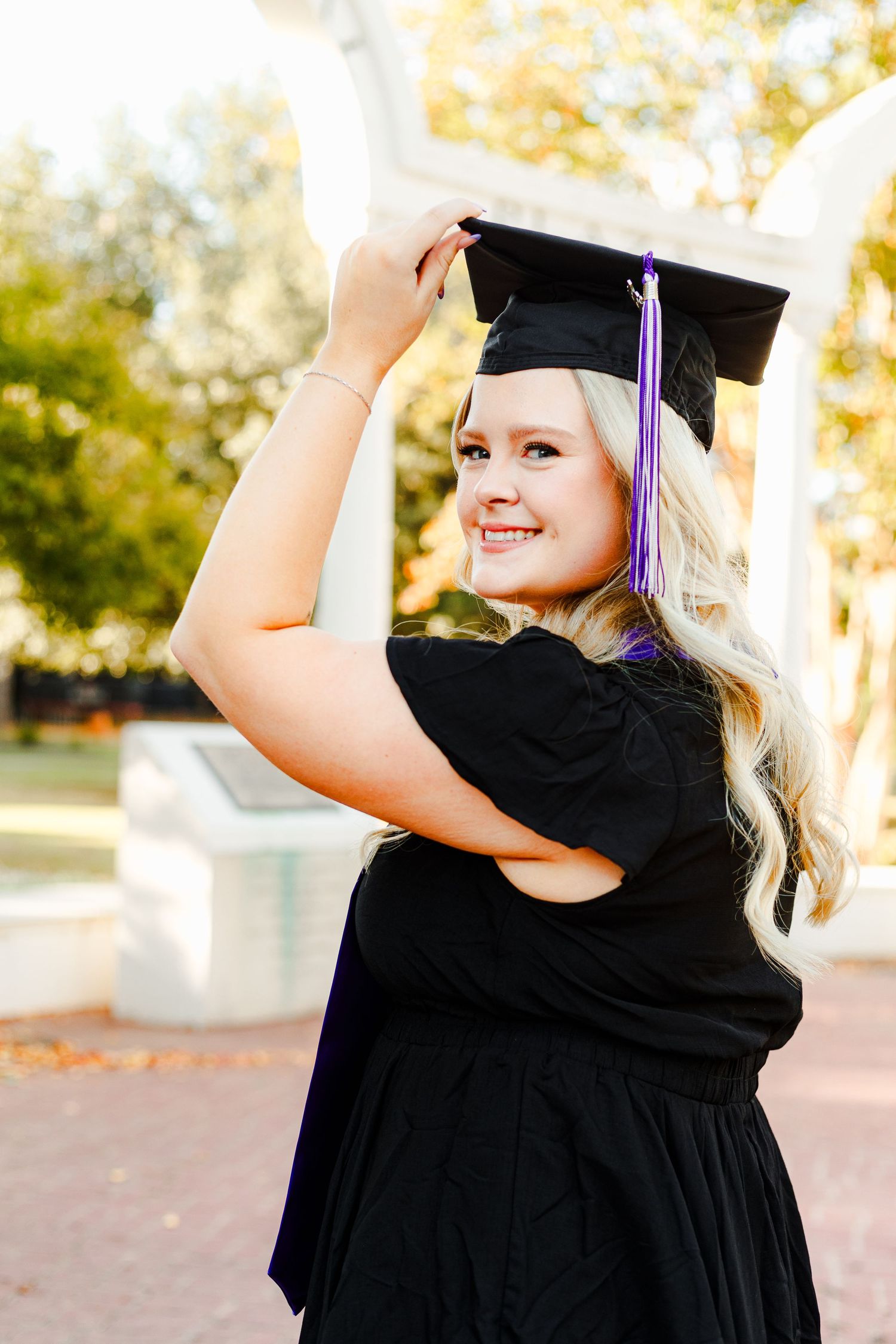 Graduate in black cap and gown smiles while touching graduation cap.