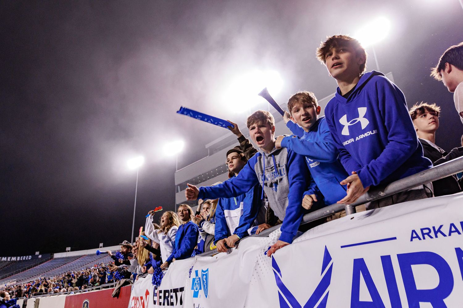 Football fans in blue jerseys cheer enthusiastically from the stadium stands during a nighttime game.