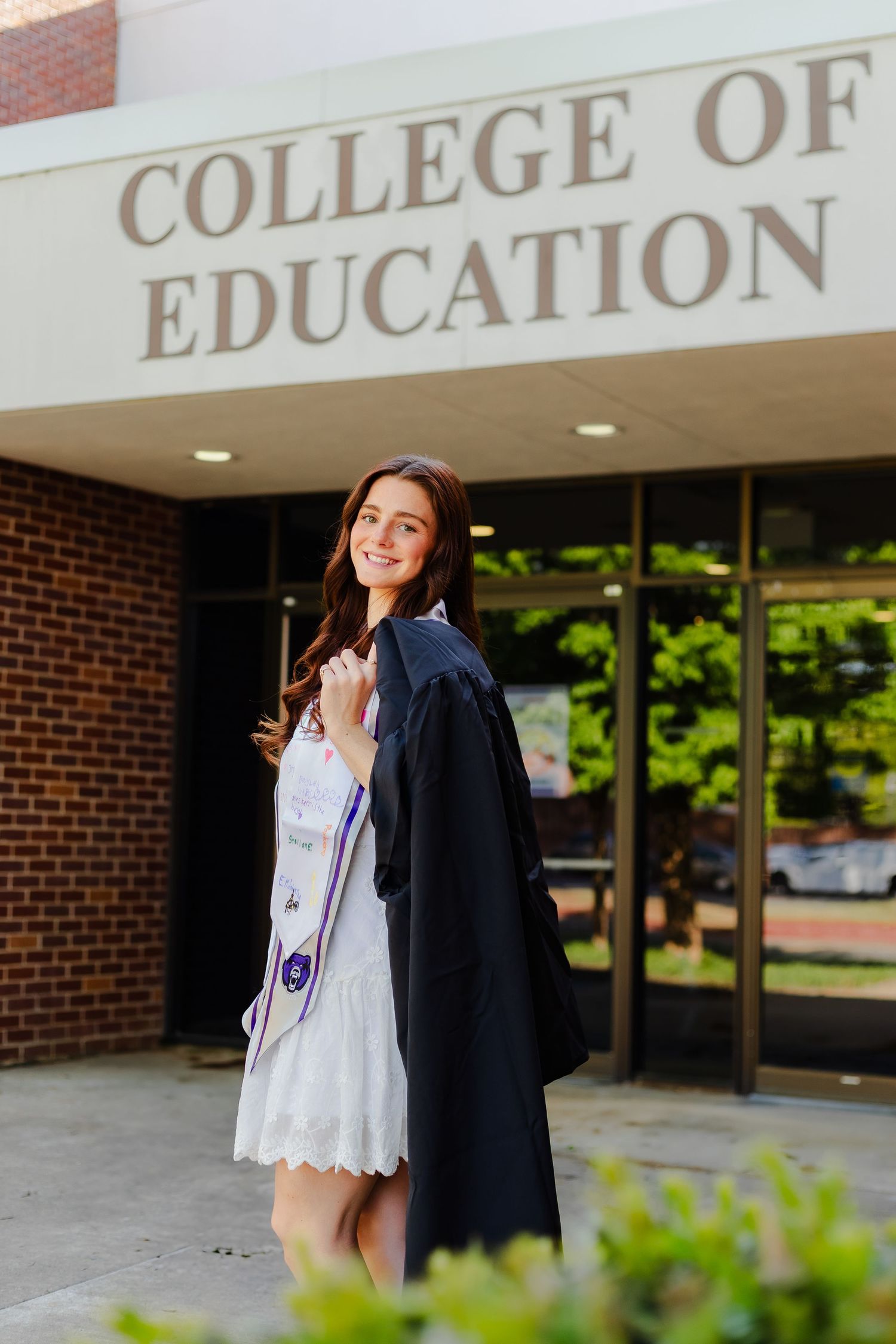 Graduate in cap and gown poses outside College of Education building entrance on graduation day.