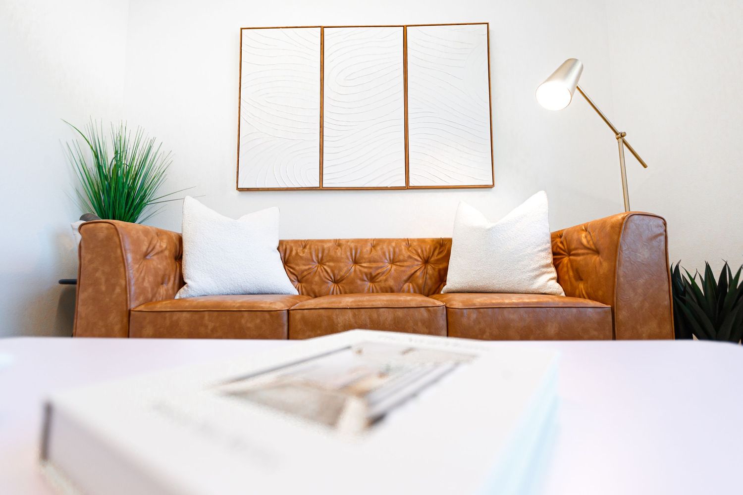 Minimalist living space featuring cognac leather sofa with white pillows and empty wooden frame art.