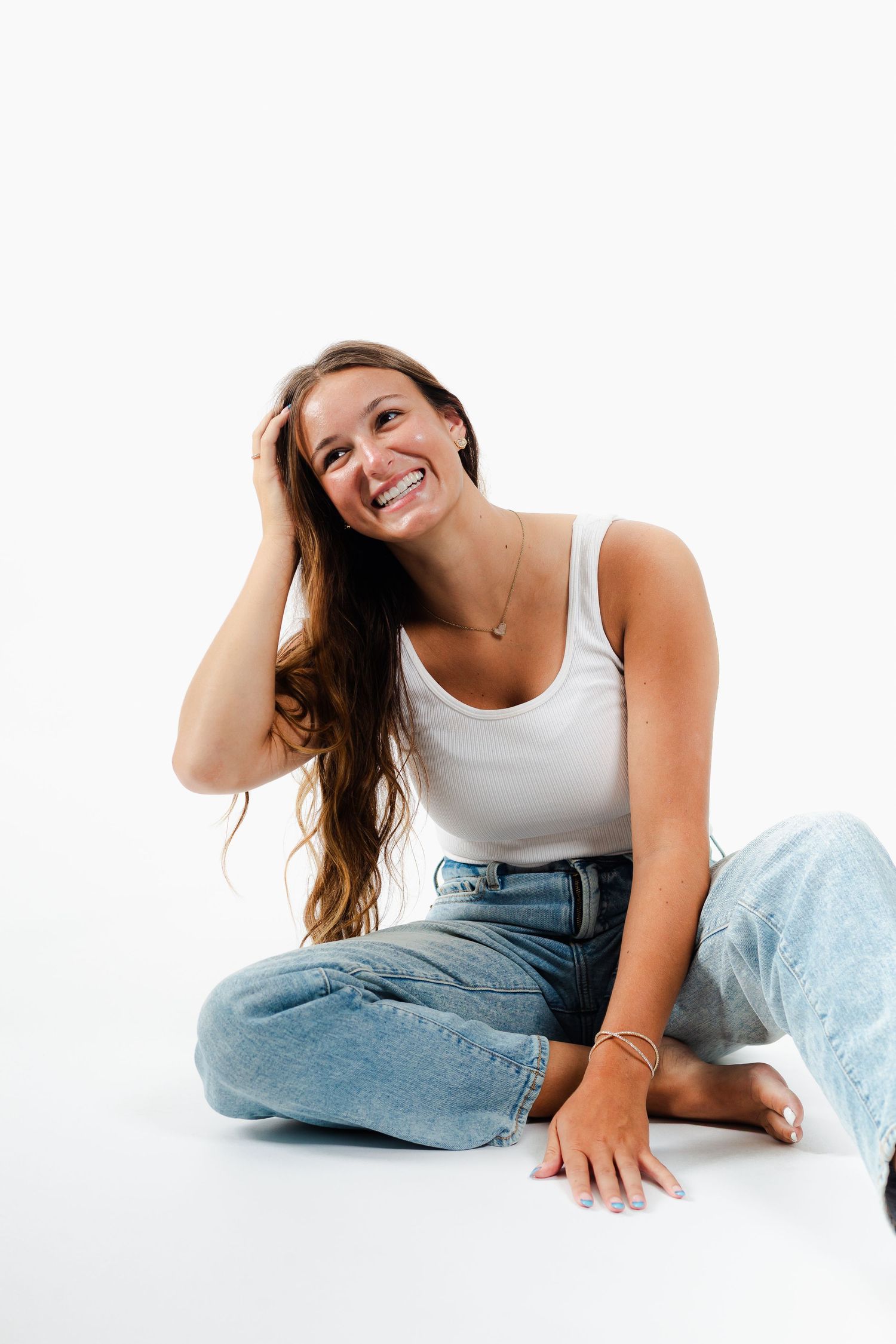Person sitting cross-legged on white floor wearing white tank top and light wash jeans while laughing joyfully.