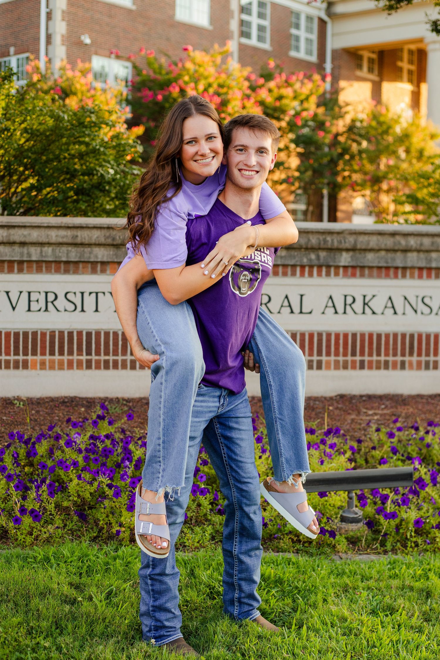Two people in purple university clothing playfully pose in front of a campus sign surrounded by colorful fall foliage.