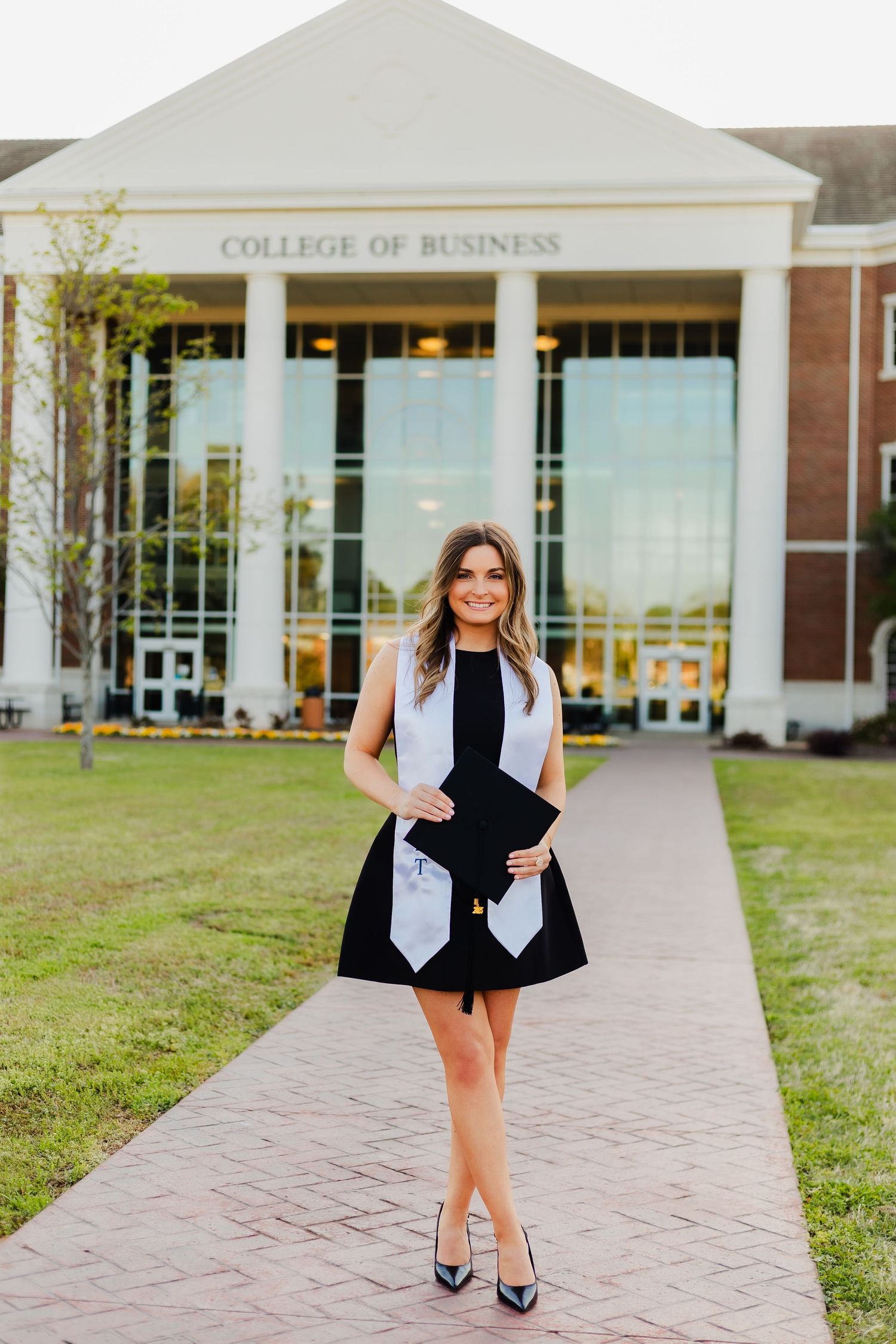 A graduating student poses in front of a College of Business building wearing a black dress and graduation stole.