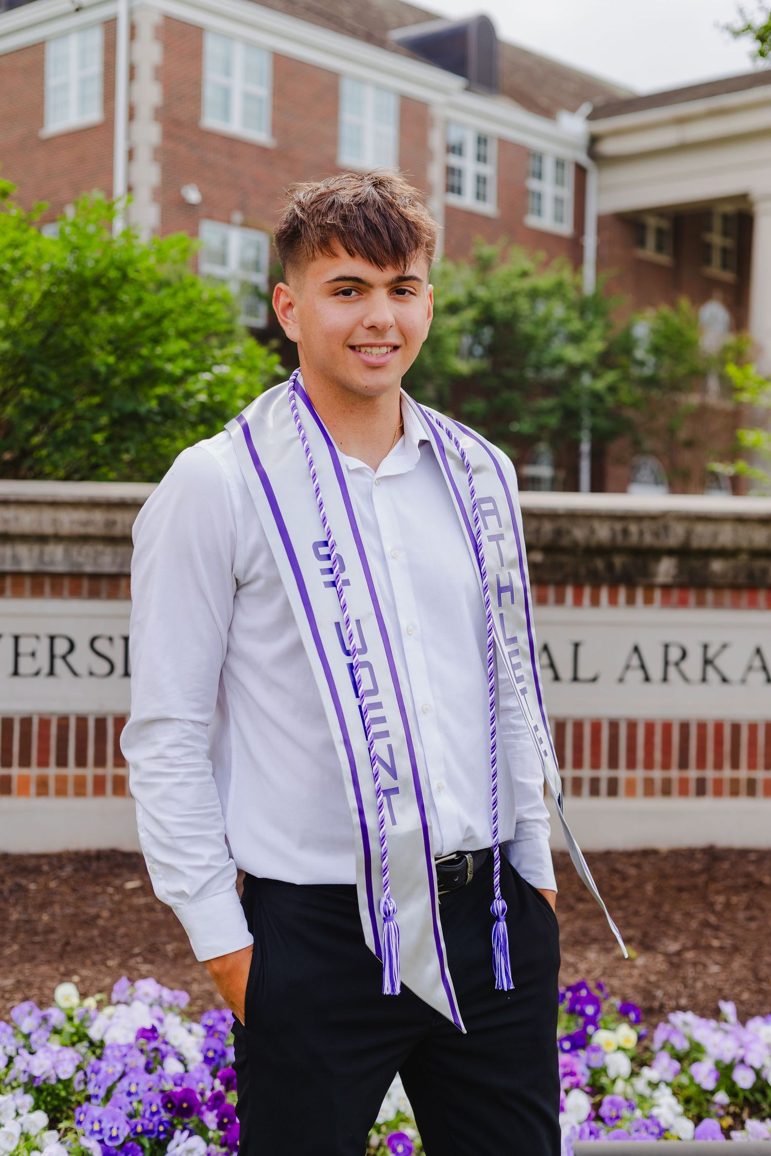 A graduate poses in front of purple flowers wearing a white shirt, black pants and purple sash at a university.