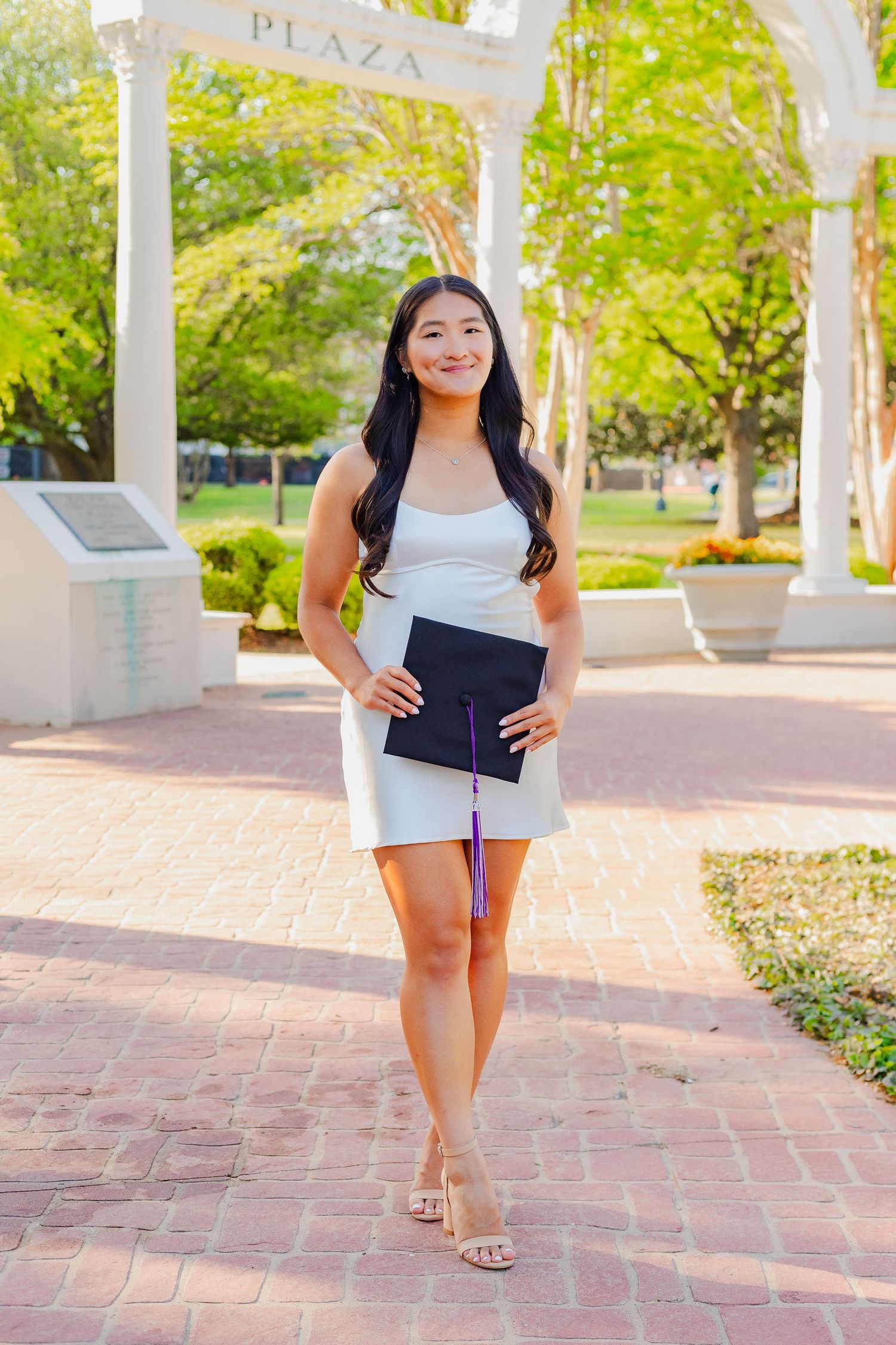 Graduate stands in a plaza wearing a white top and dark bottom while holding a diploma in an outdoor setting.