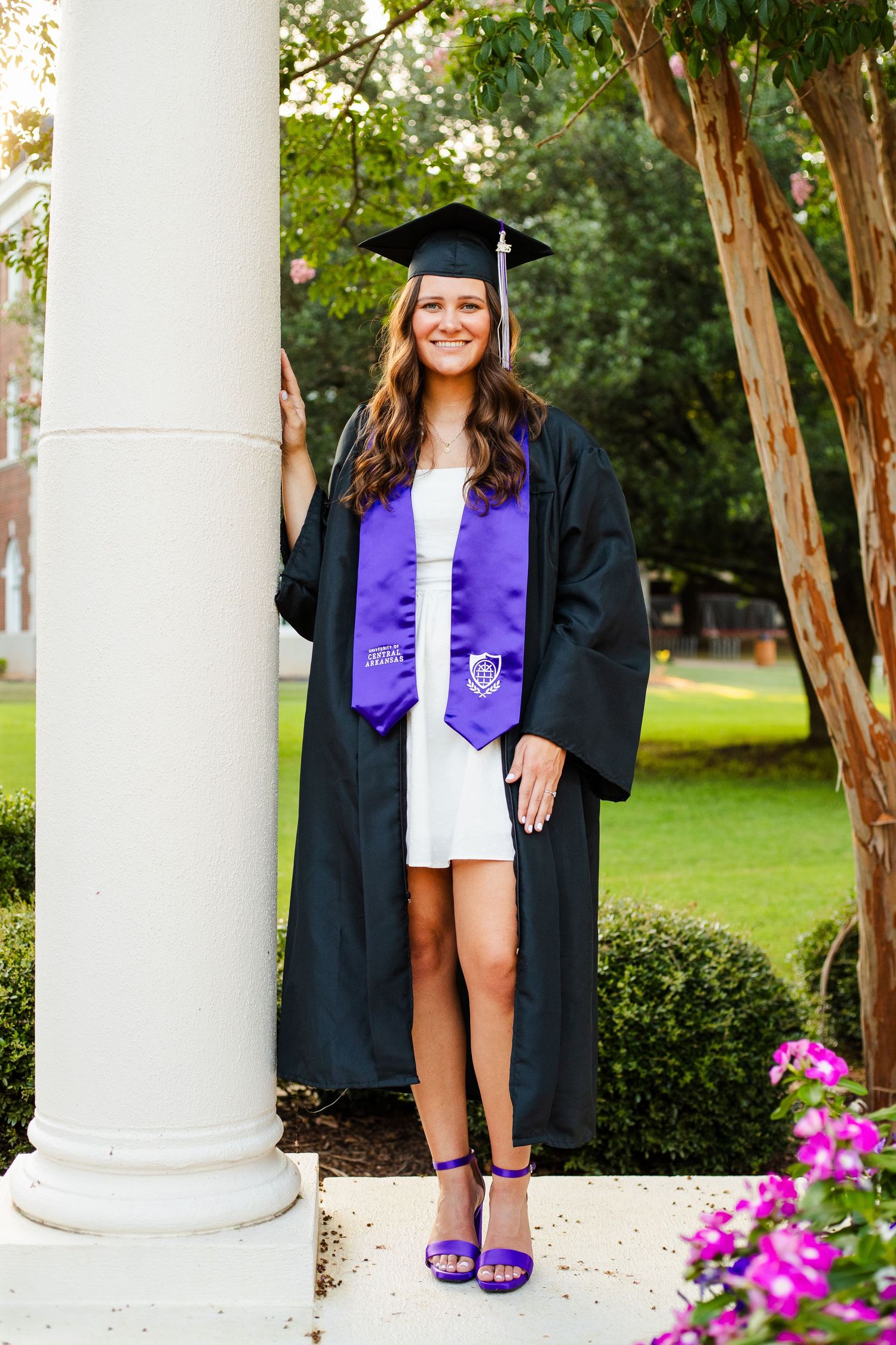 Graduate in cap and gown with purple stole and graduation sash poses by white column with flowers in background.
