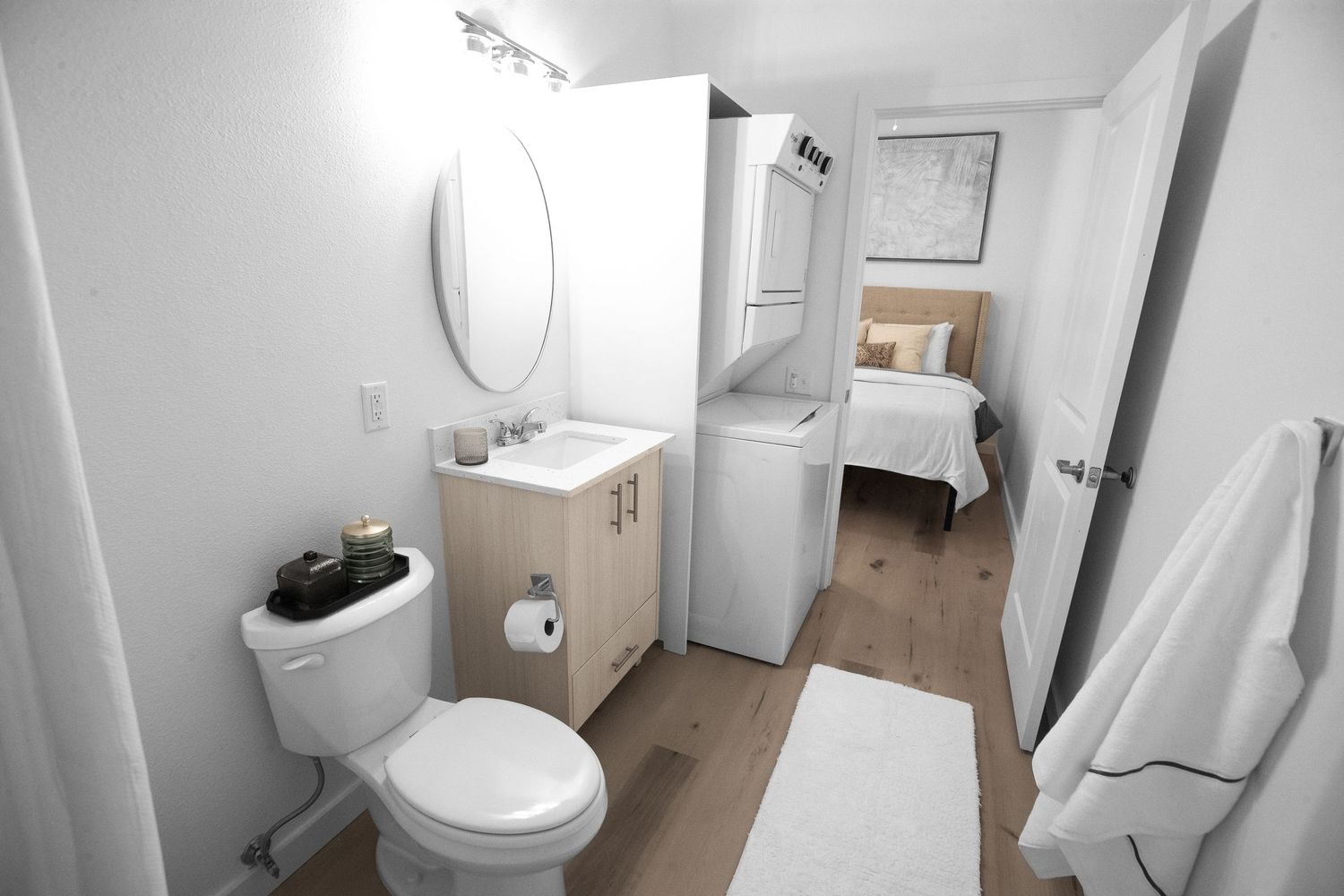Compact bathroom with white vanity, round mirror, and visible bedroom through doorway.
