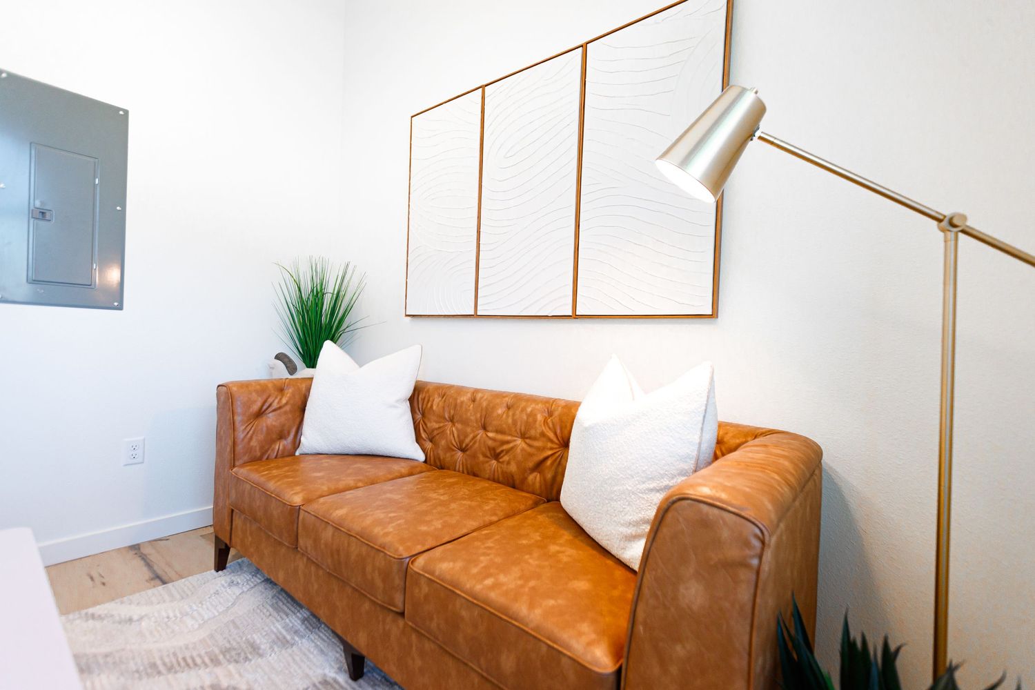 Modern living room with tan leather couch, white throw pillows, wooden frame art, and brass floor lamp.